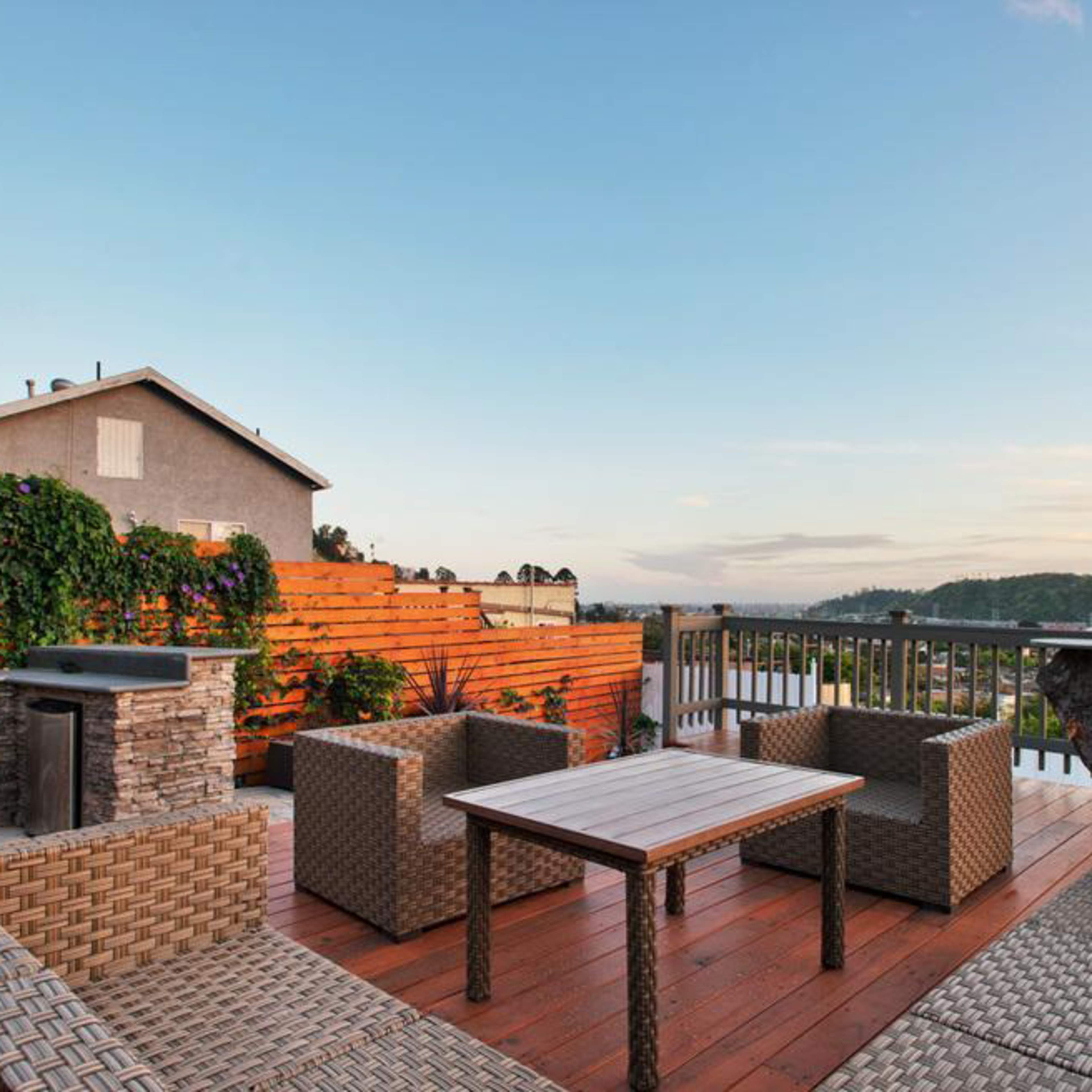A wooden deck with a table and chairs, a grill, and a hillside view in the background.