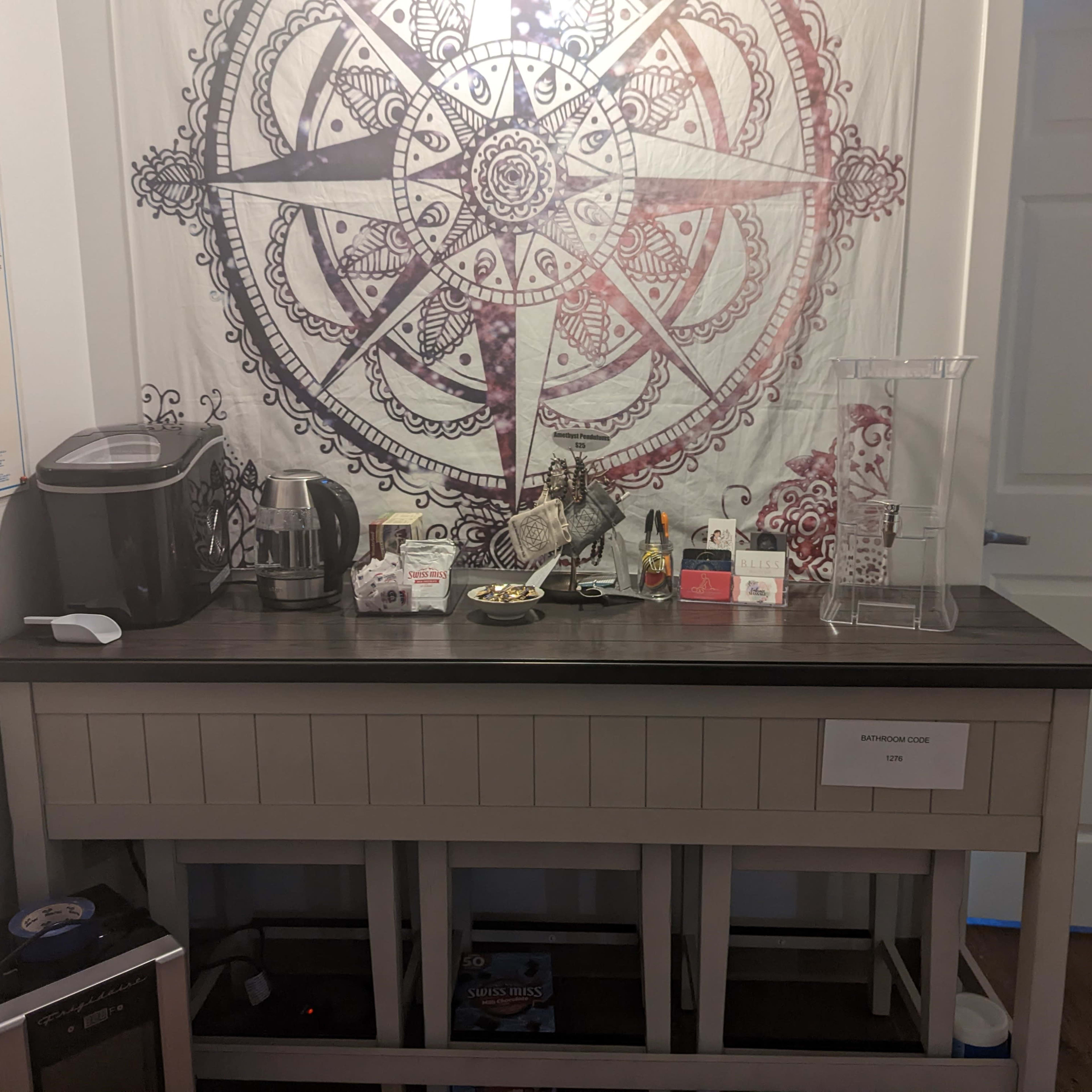 A room corner with a table displaying a coffee machine, toaster, snacks, and decorative items against a wall featuring a large mandala tapestry.