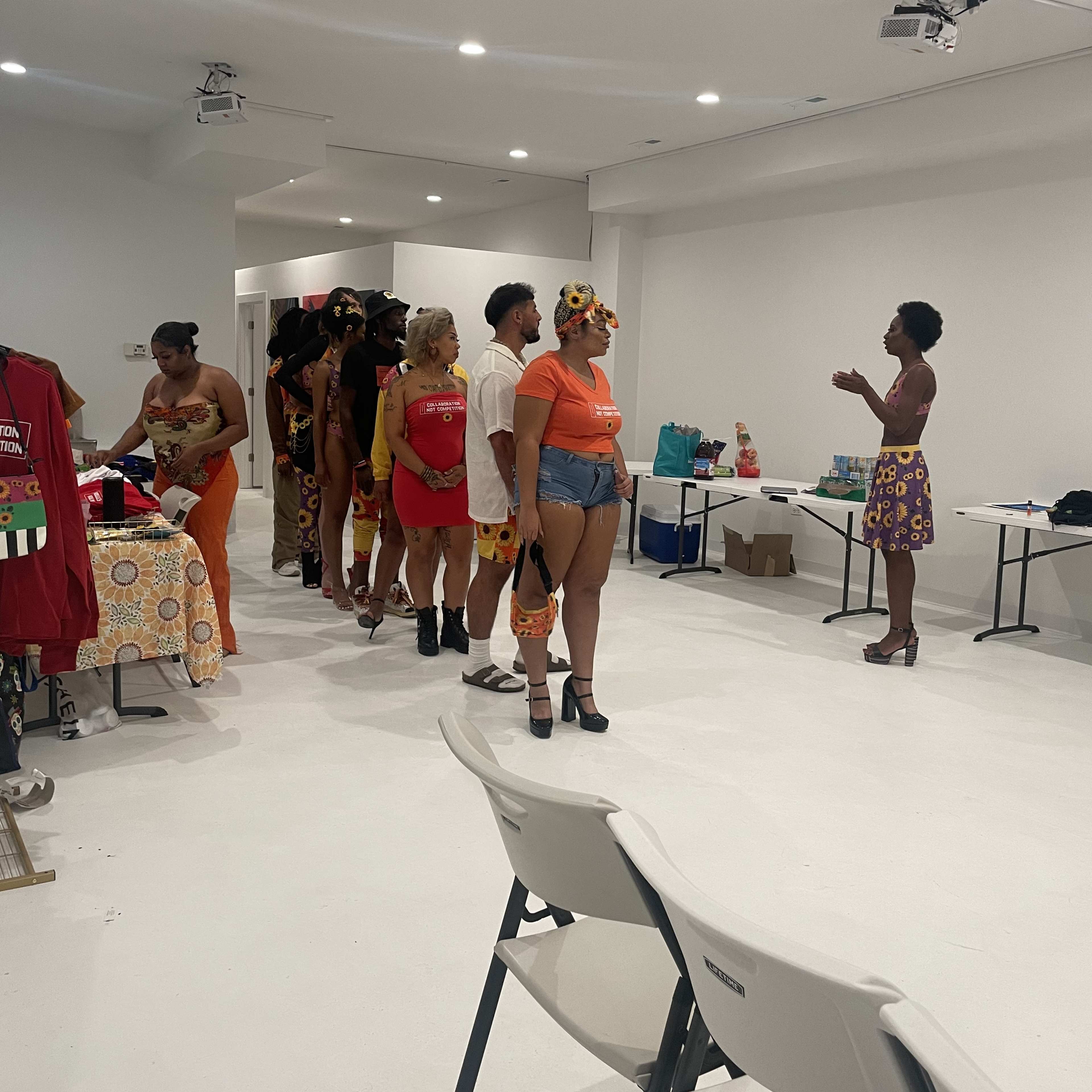 A line of people dressed in colorful outfits stands in an indoor space, while a person gestures towards them from the front.