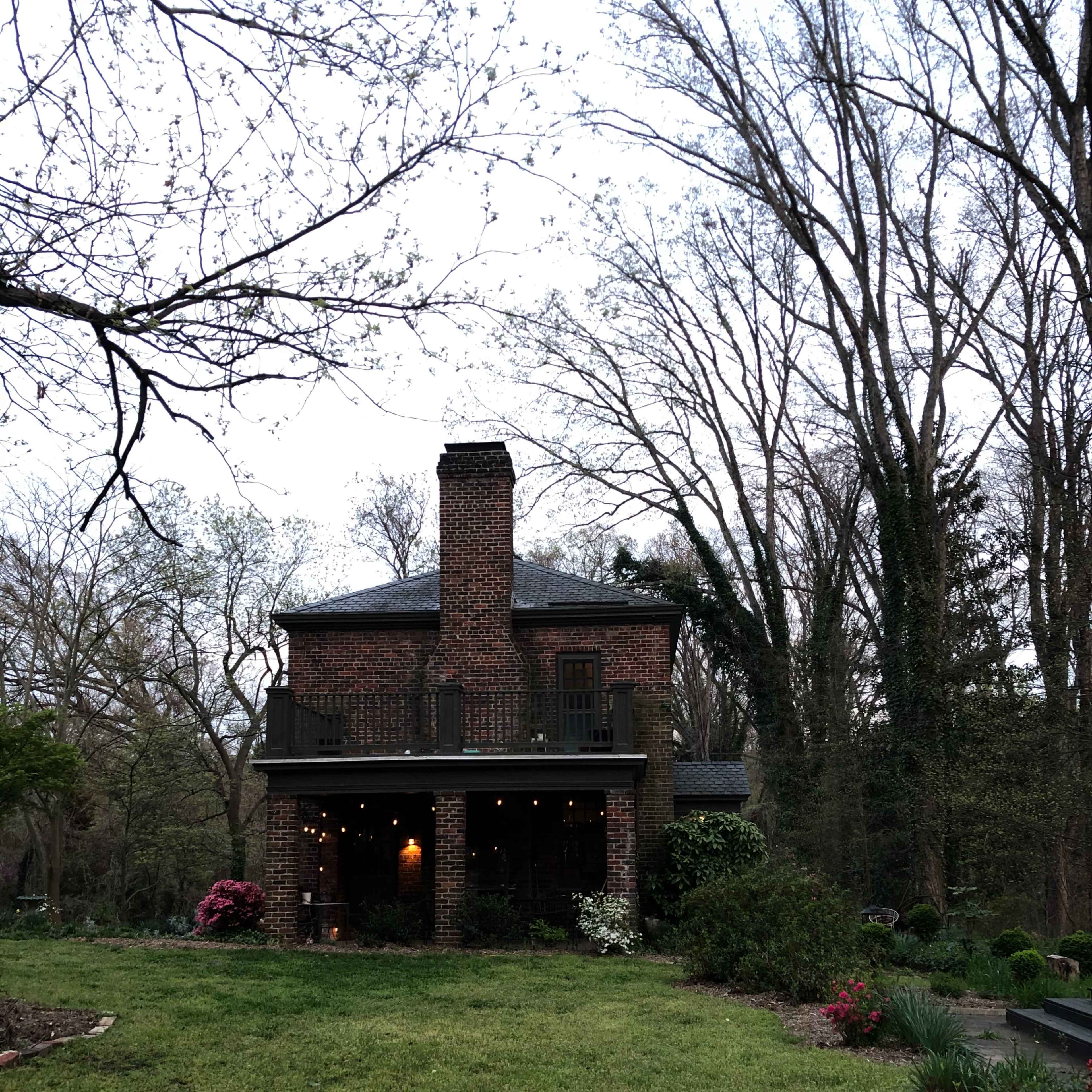 A brick house with a chimney and a porch is surrounded by trees and shrubs in a landscaped yard.