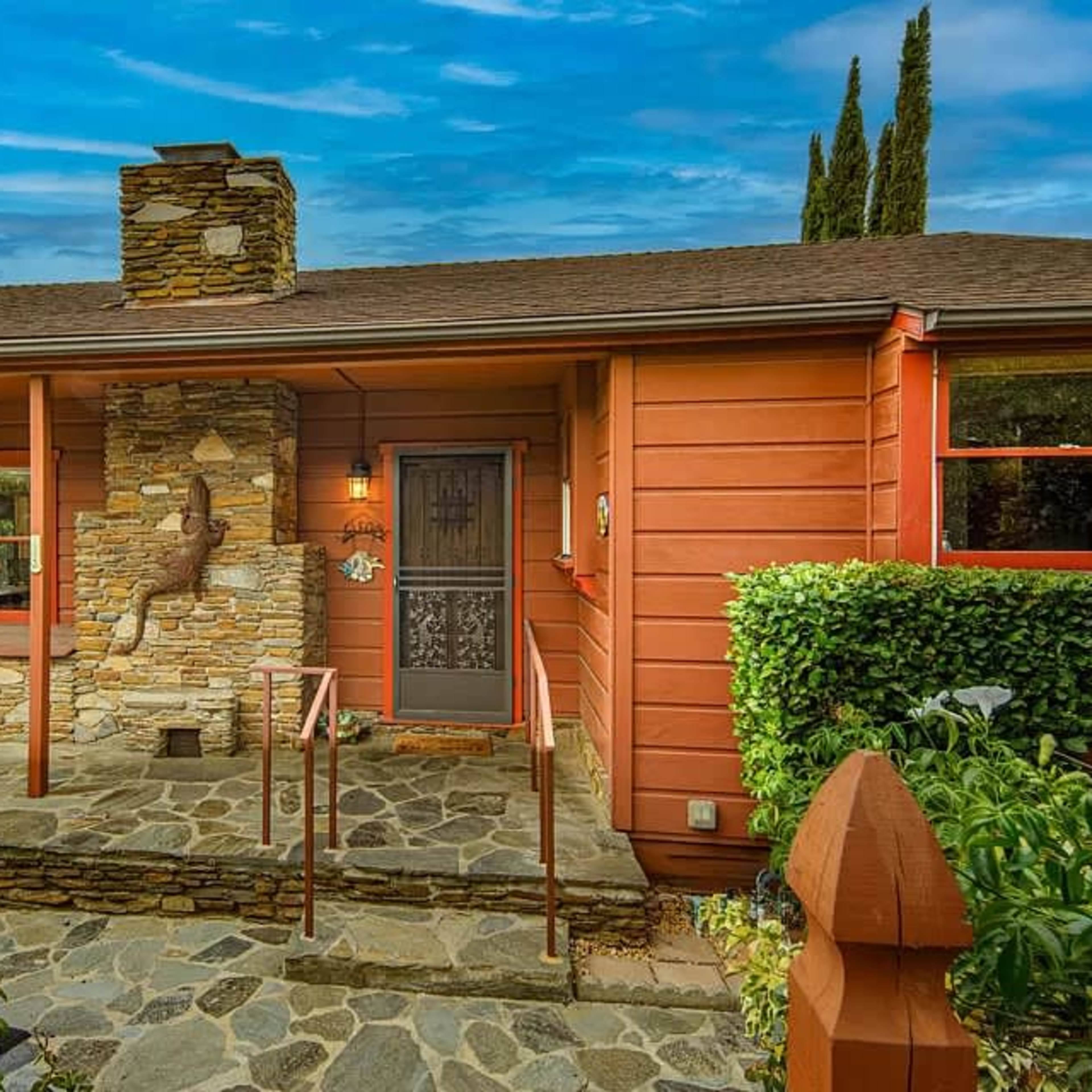 A single-story wooden house with red siding, a stone chimney, and a stone pathway leading to the front door, surrounded by greenery.