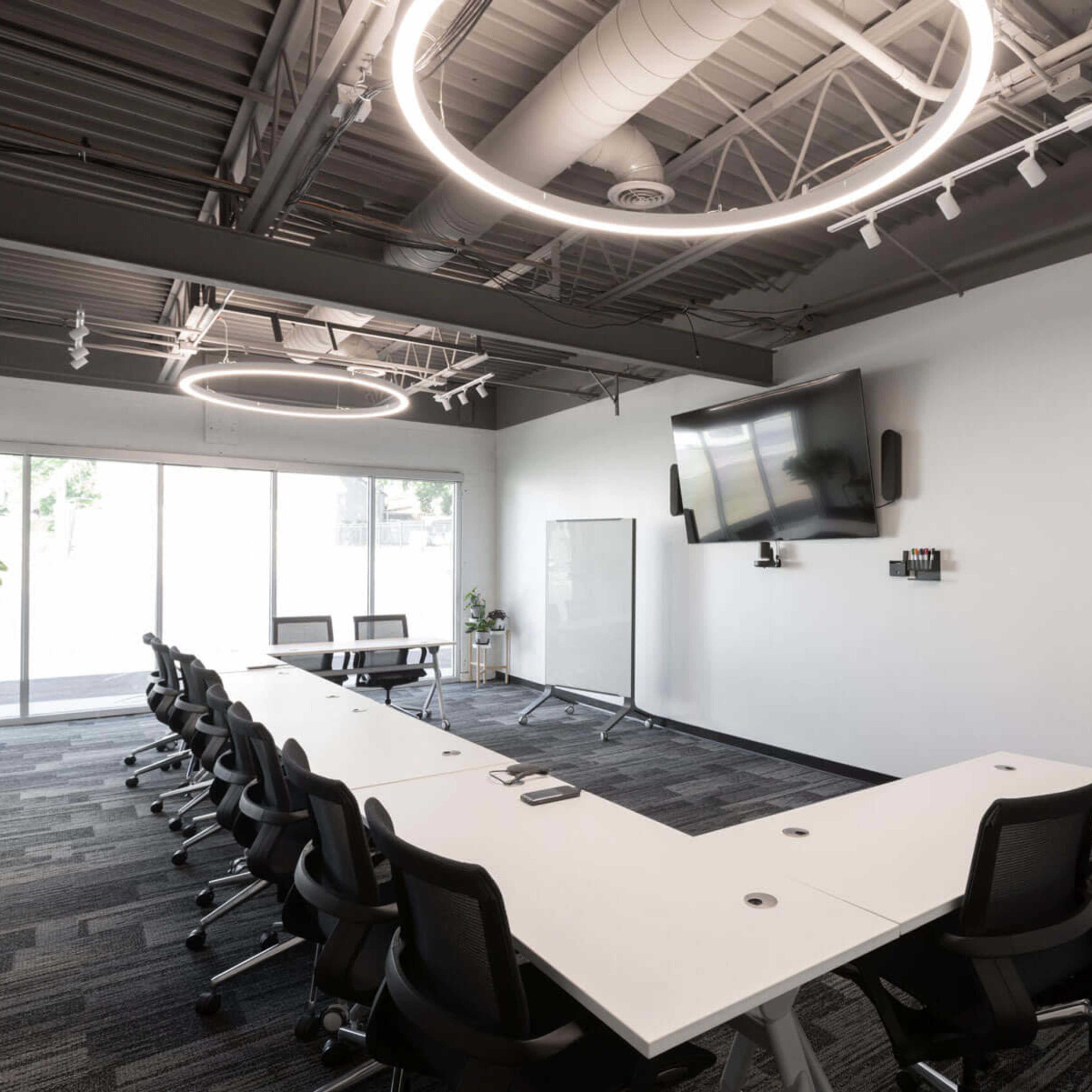 A modern conference room with a long, white table surrounded by black chairs, large windows, and circular light fixtures overhead.