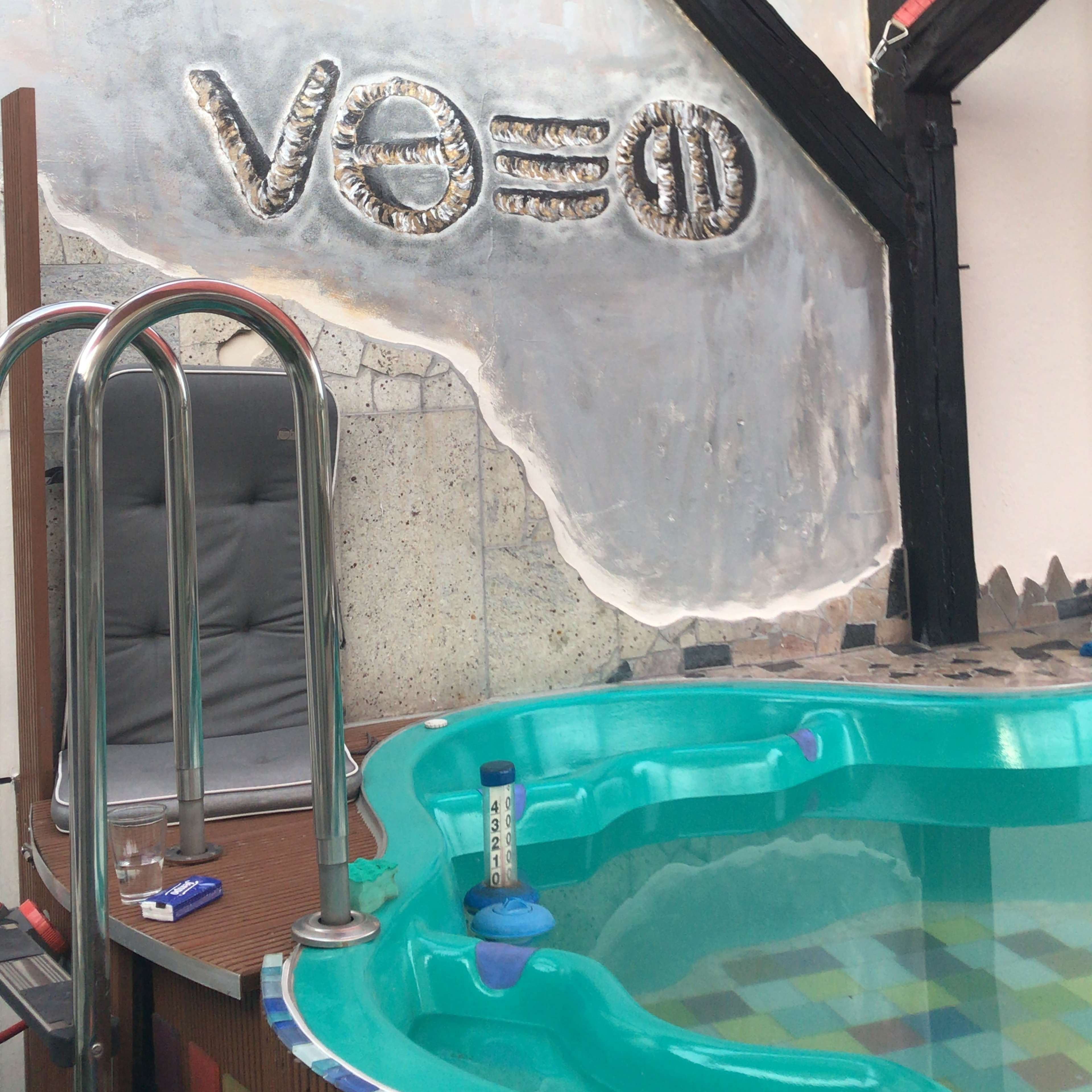 A turquoise hot tub with a partially finished wall featuring metallic letters and symbols in a shaded area.