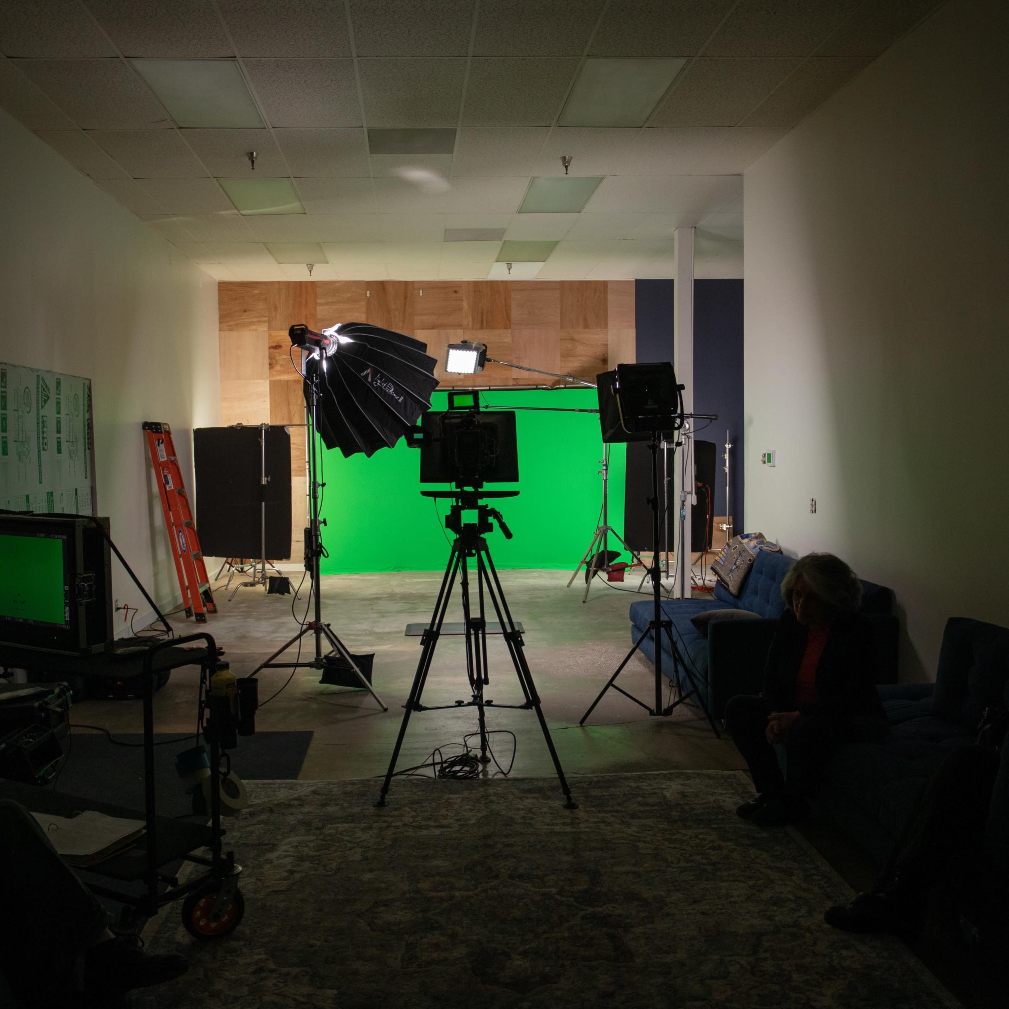 A film studio setup featuring a green screen, lighting equipment, and several crew members seated on couches.
