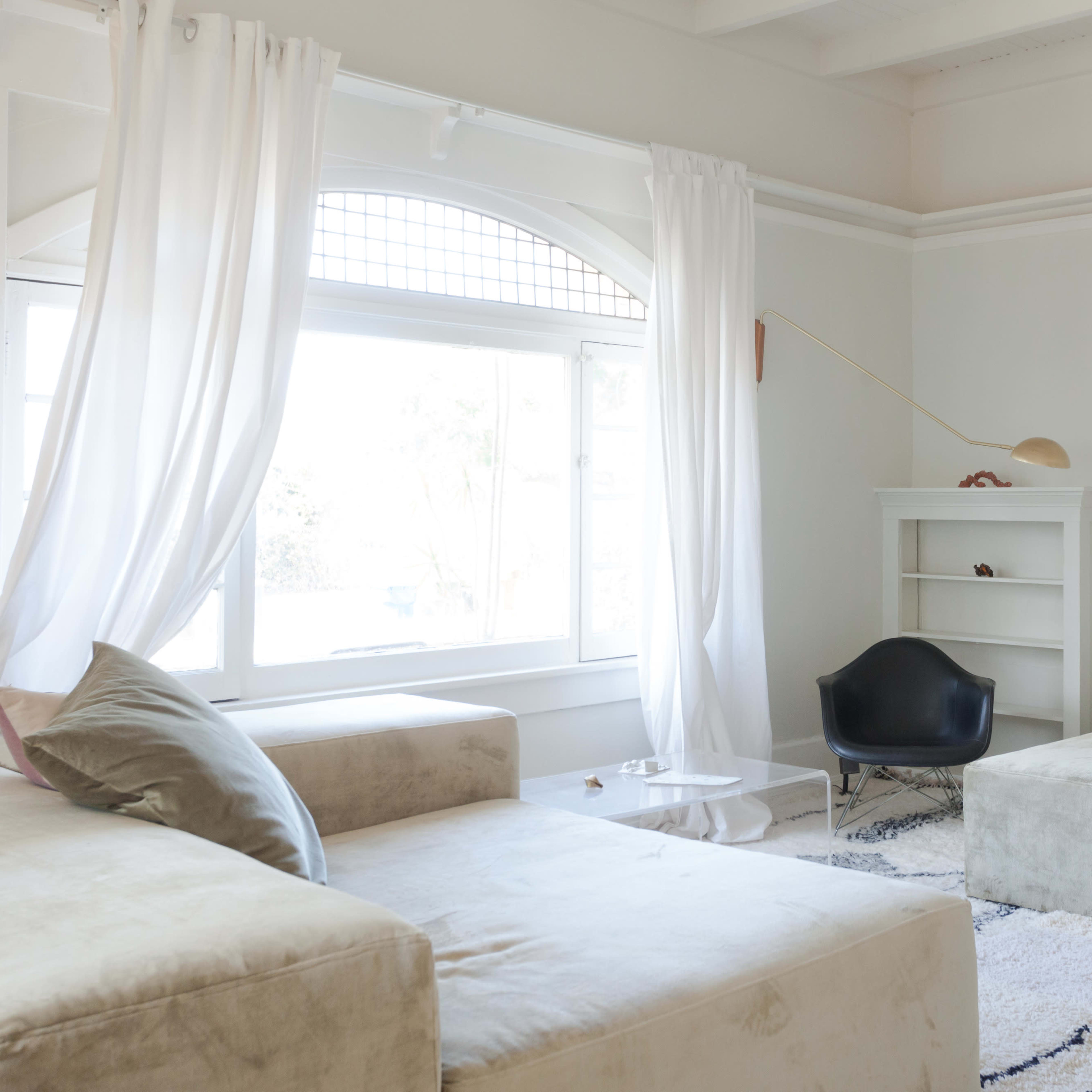 A bright living room features a large window, minimalist furniture, and sheer curtains, creating a spacious atmosphere.