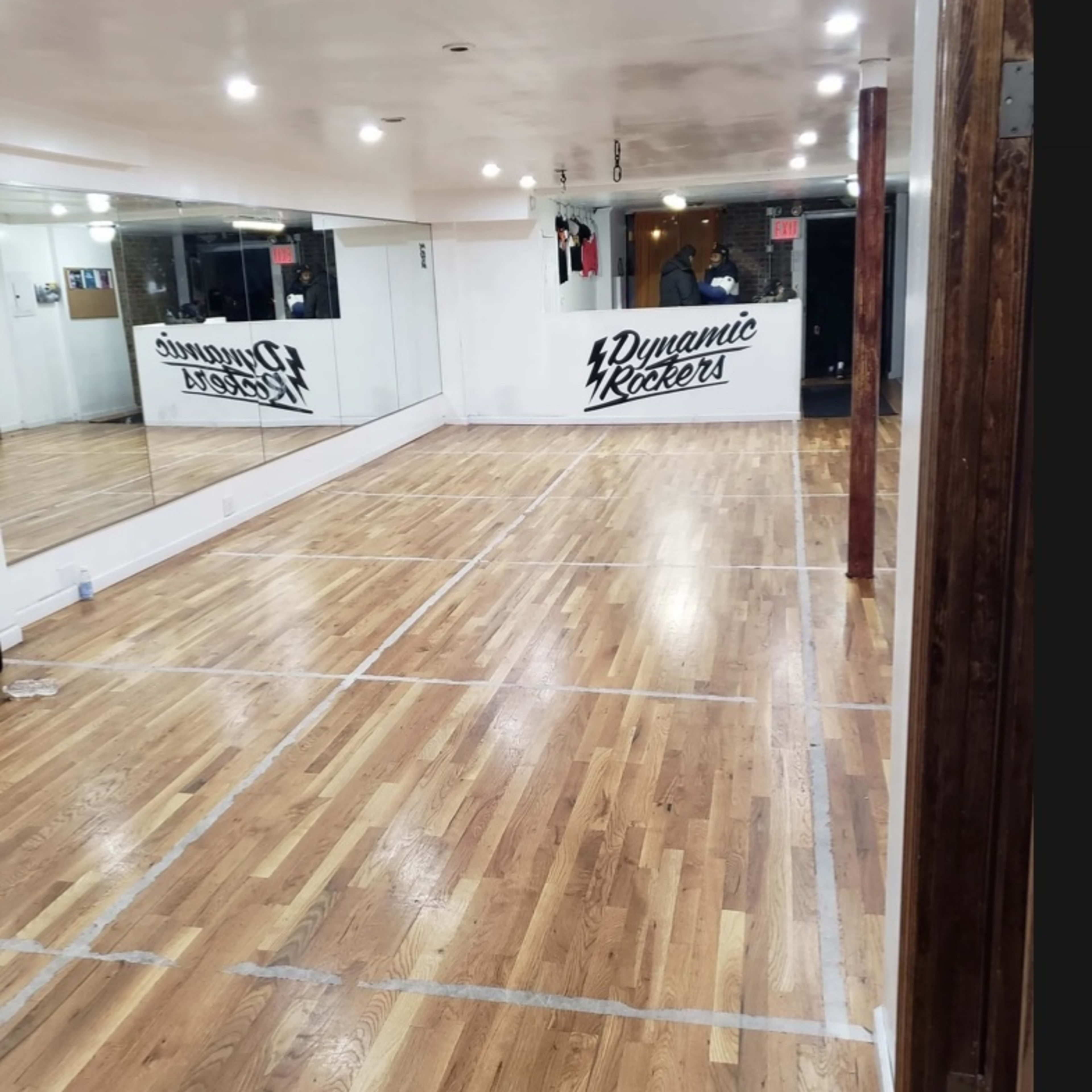 An empty dance studio with polished wooden floors and a mirror on one wall, featuring the name "Dynamic Rockers" painted on another wall.