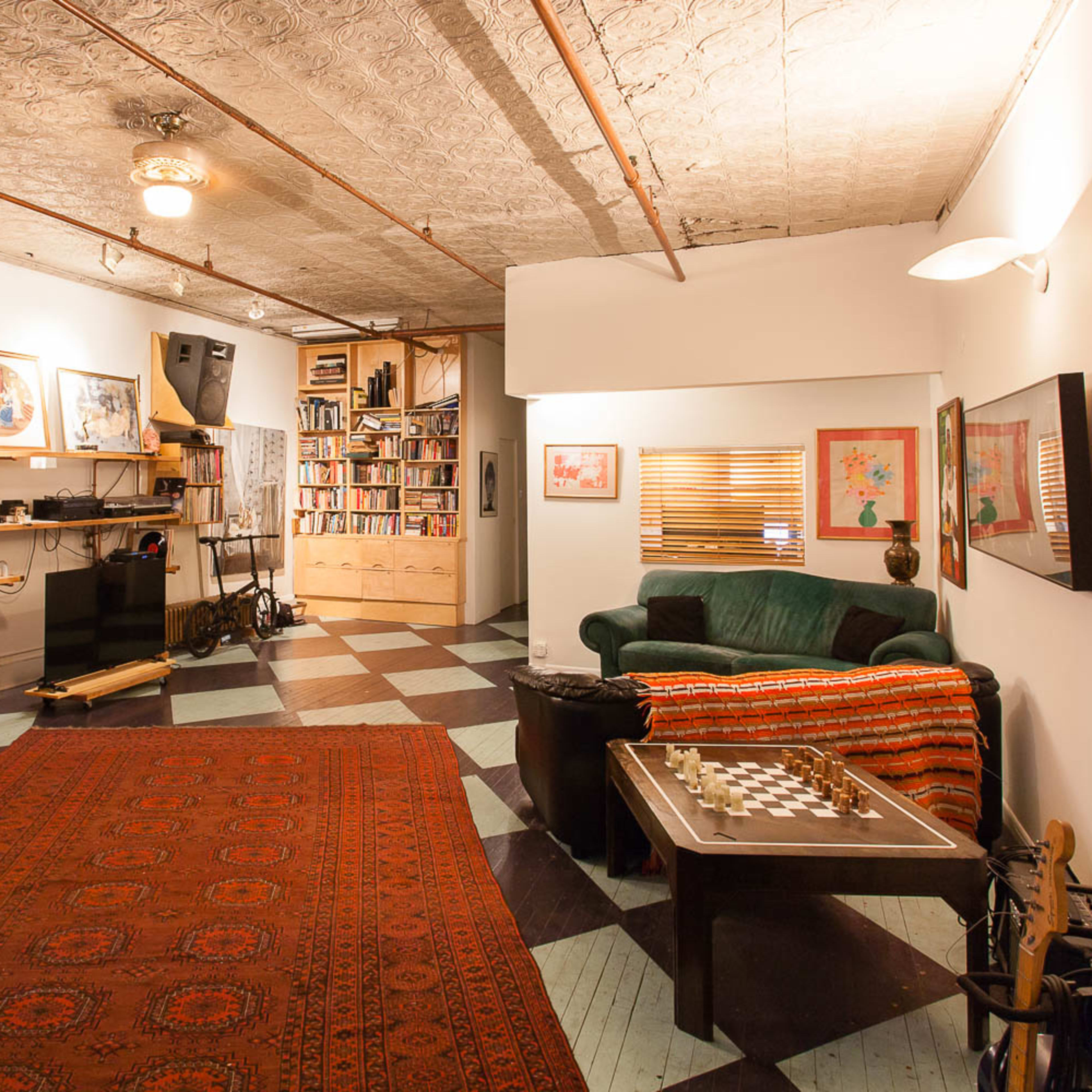 A spacious room features a green couch, a chess table, shelves filled with books, large speakers, and various musical instruments against patterned flooring.