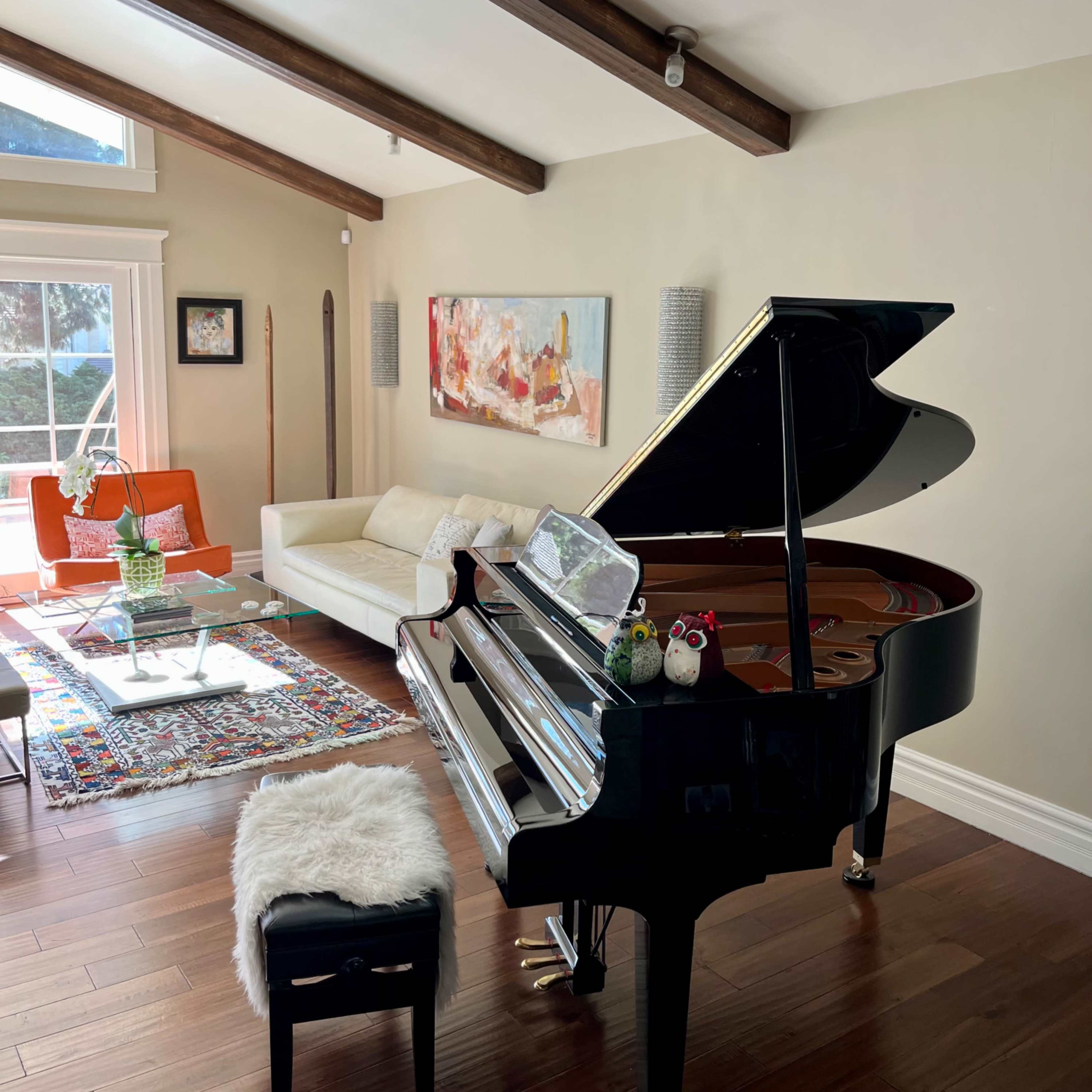 A grand piano sits in a sunlit living room featuring a white sofa, vibrant artwork, and an area rug.