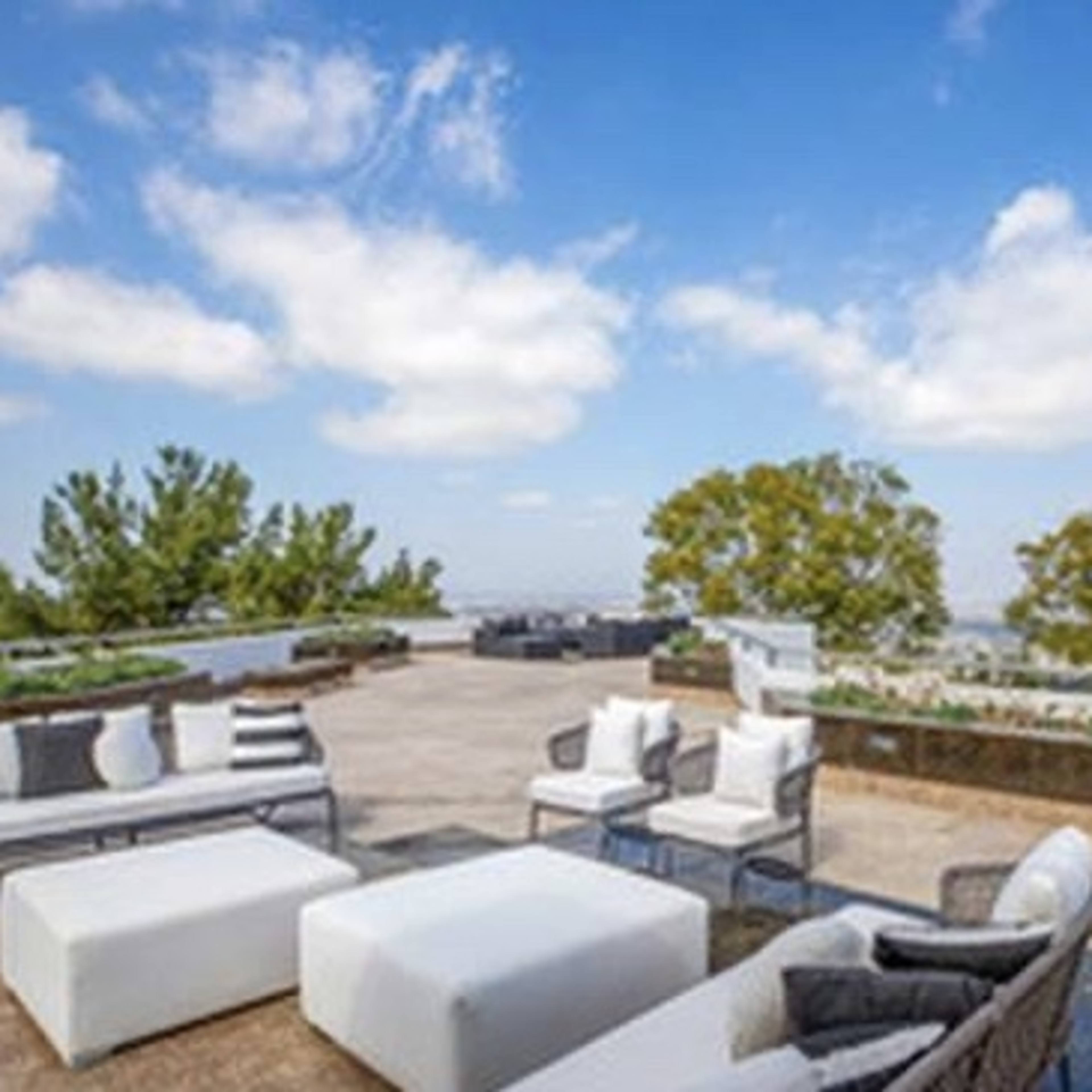A modern outdoor terrace features several seating arrangements, a low table, and a panoramic view of the landscape under a blue sky with scattered clouds.
