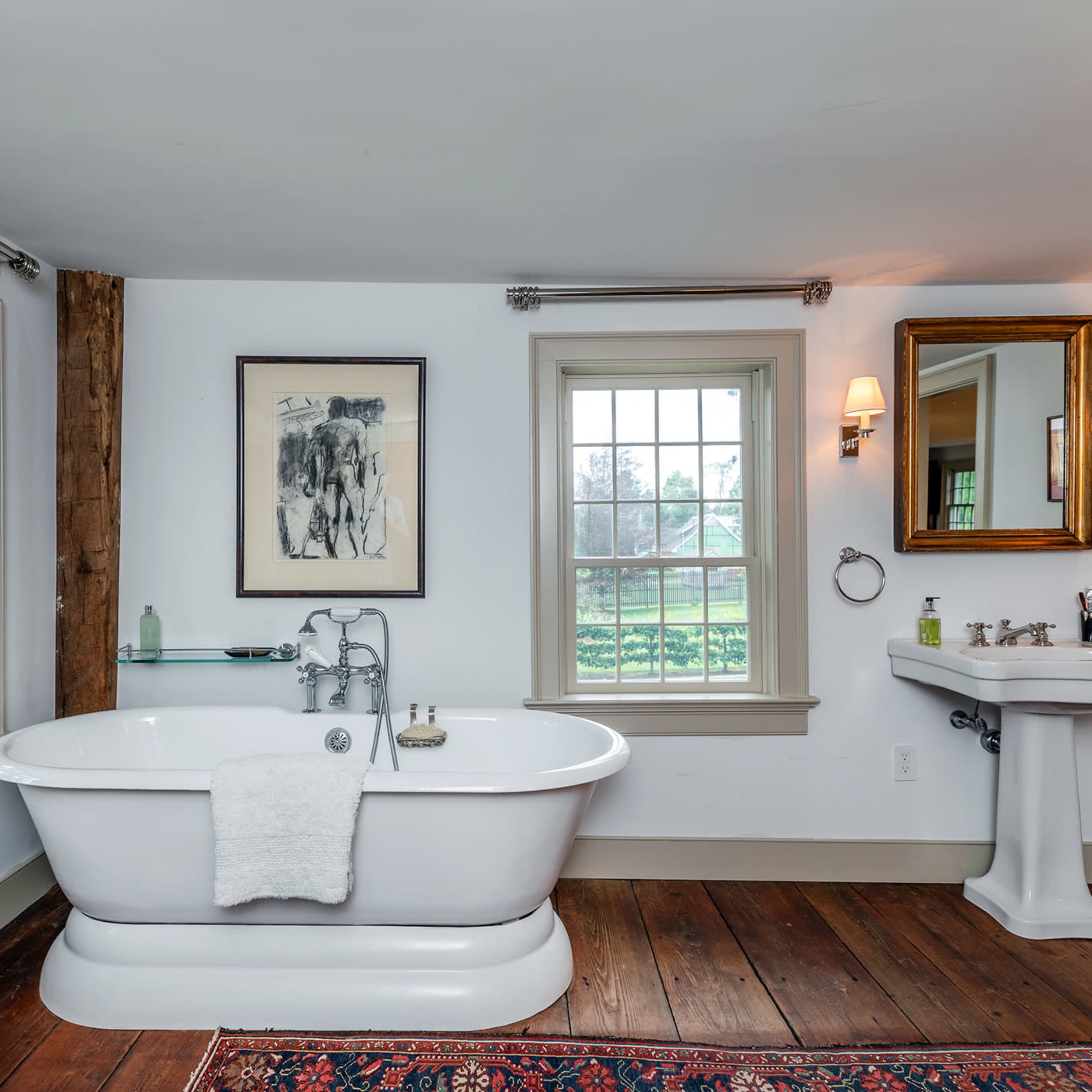 A bathroom featuring a vintage freestanding bathtub, a pedestal sink, wooden flooring, and large windows with views of greenery.