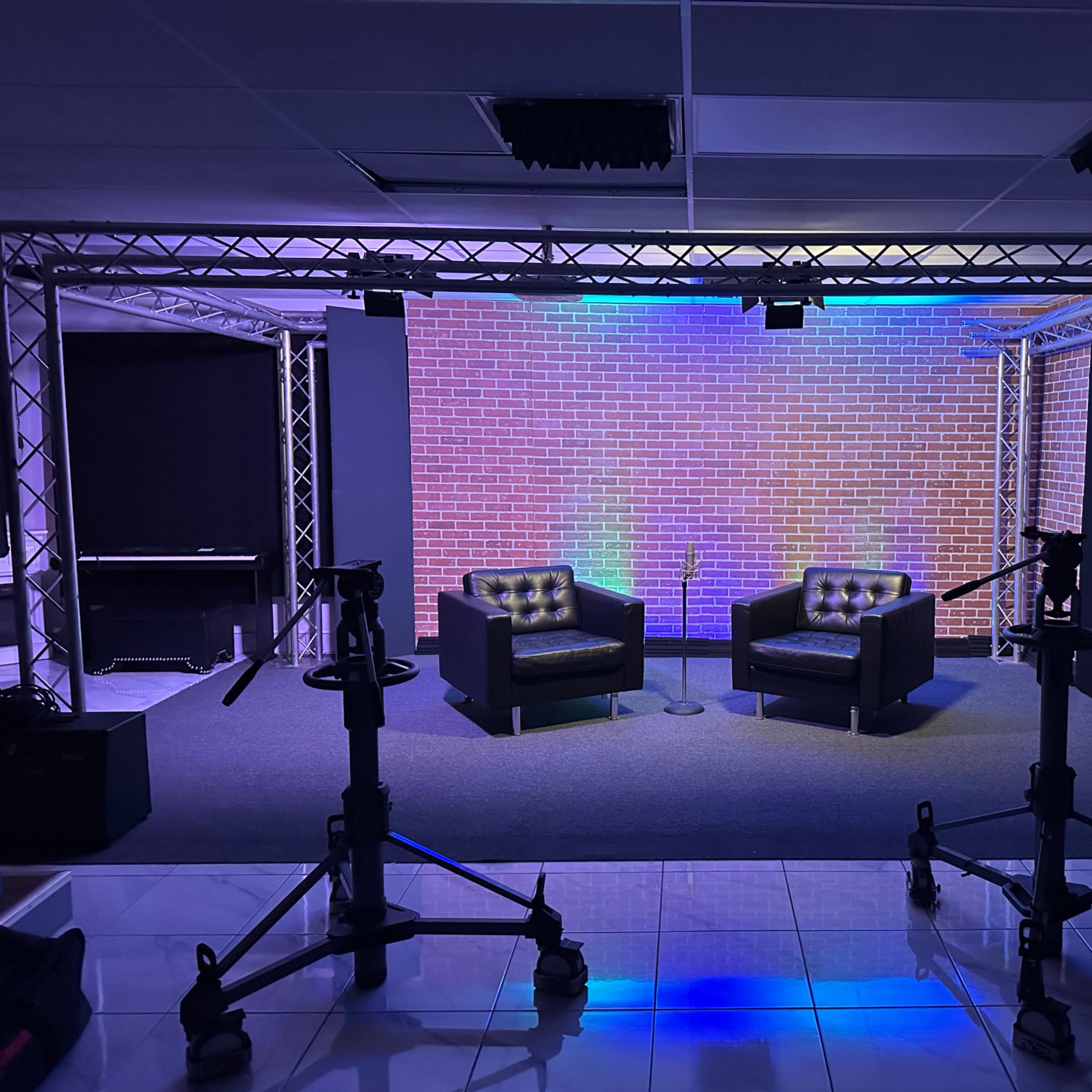 A staged interview setup with two black upholstered chairs, a brick wall backdrop, and lighting on a gray carpet, surrounded by equipment stands.