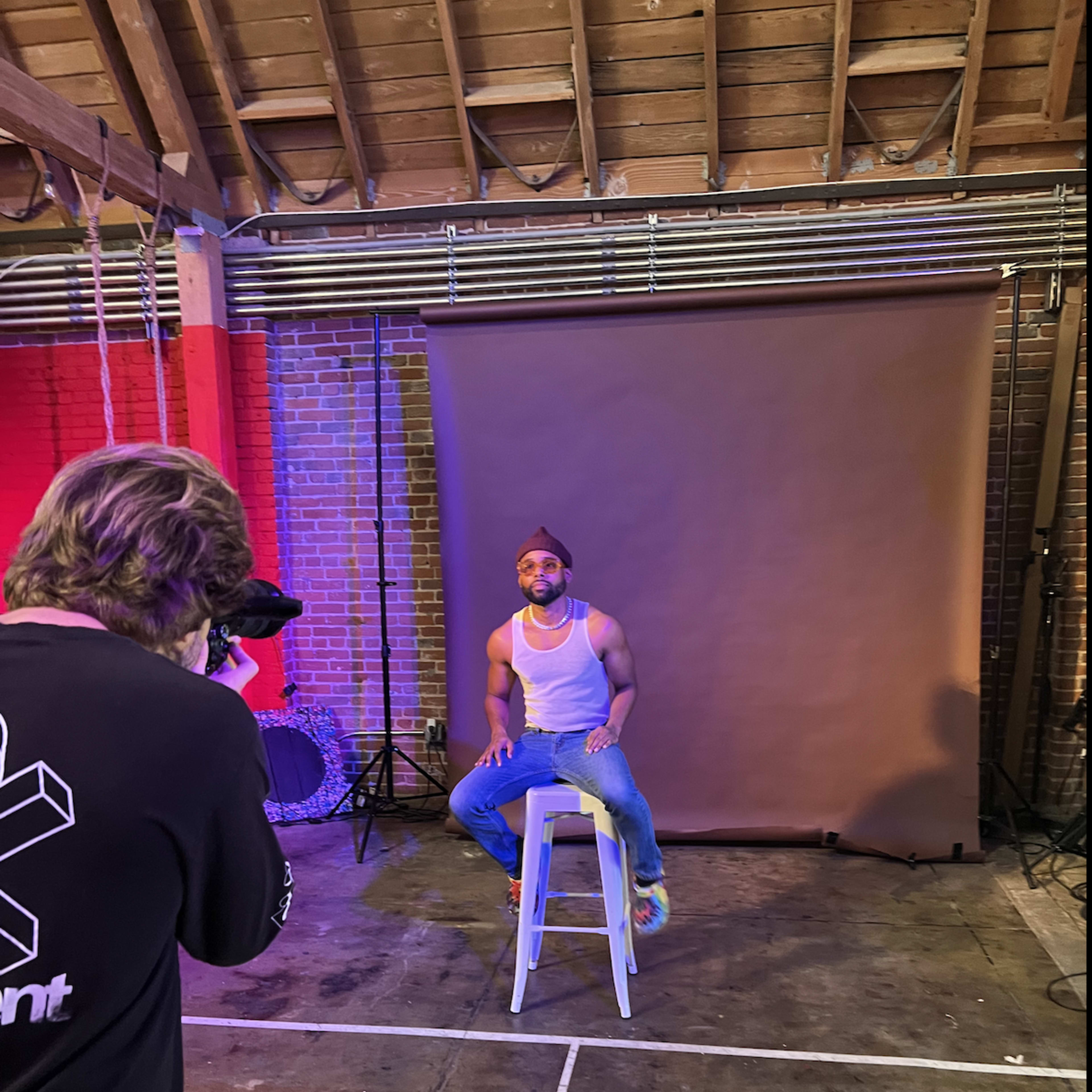 A person is sitting on a stool in front of a backdrop, while another person is taking photos with a camera.