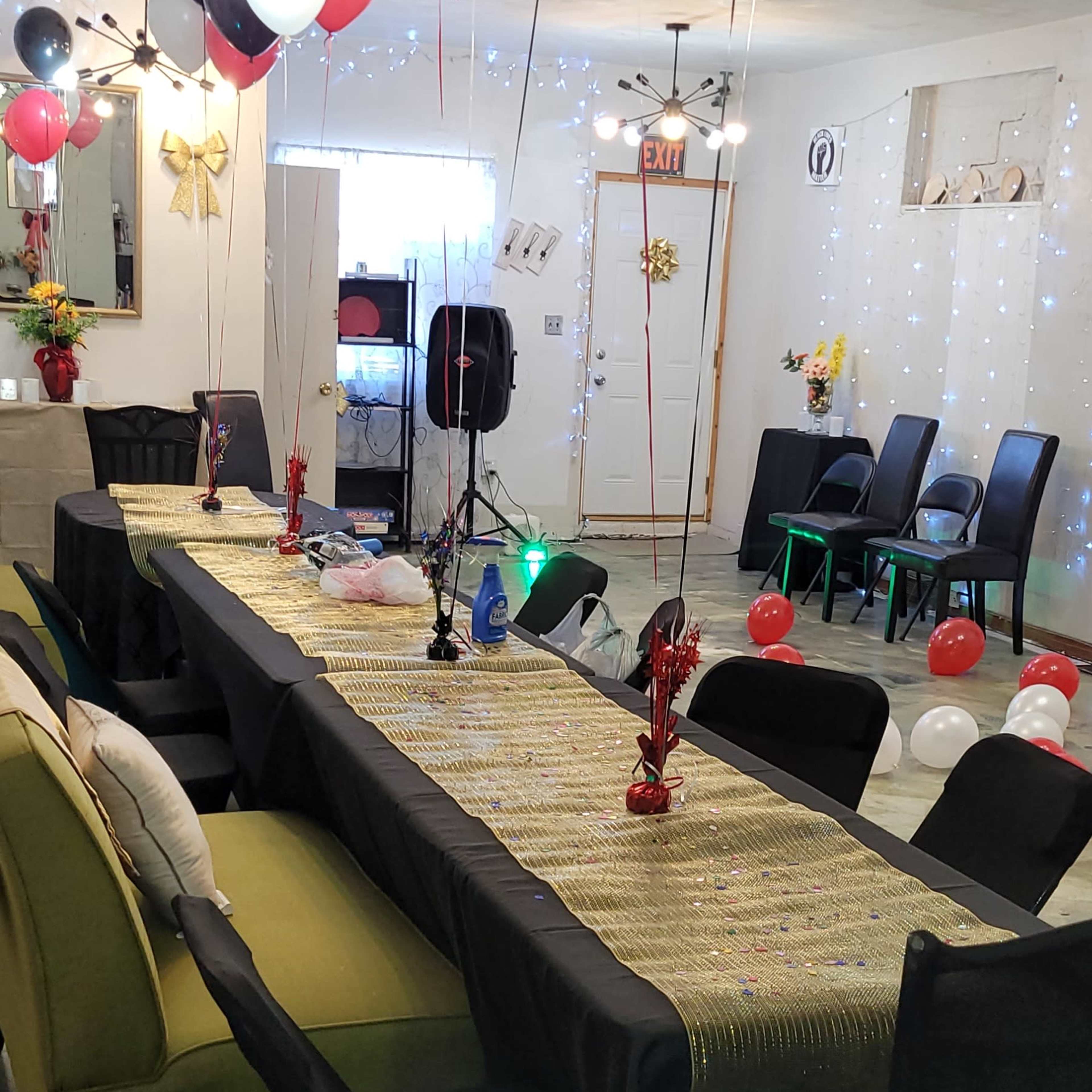 A decorated party space featuring a long table with a gold table runner, black and red balloons, and chairs arranged around the area.