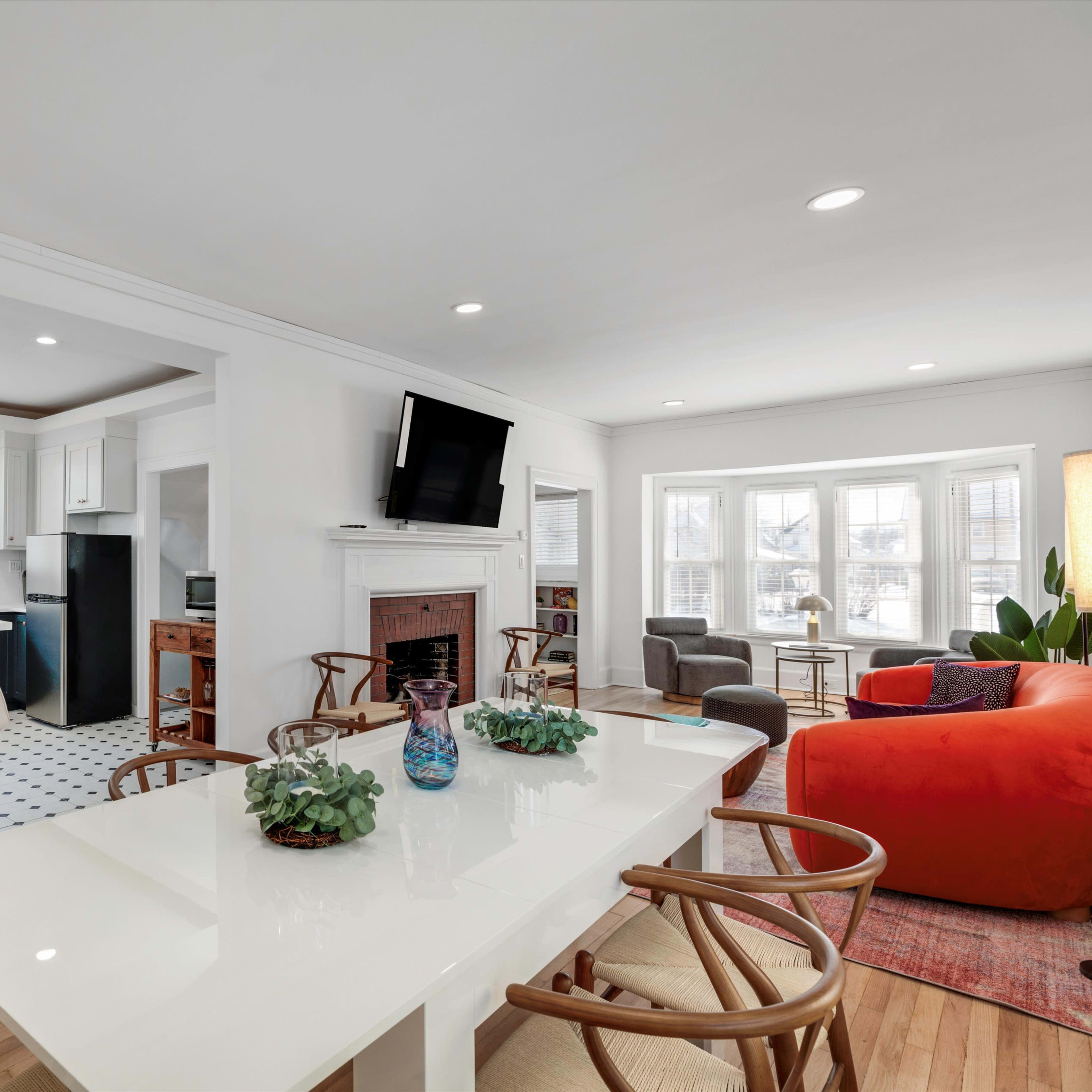 A modern kitchen and dining area features a white table, bar stools, and a living room with a large window and colorful furniture.