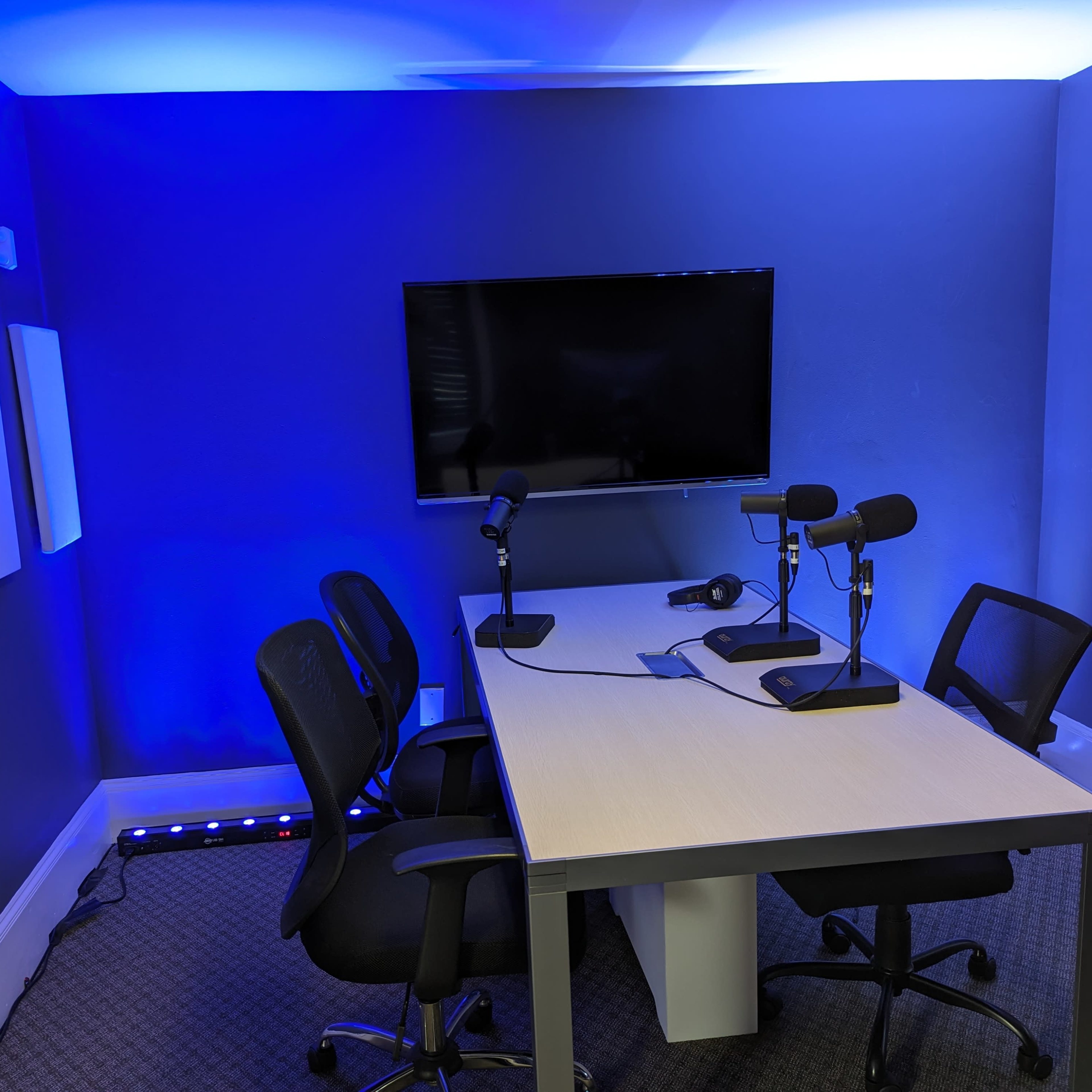 A recording studio features a table with two microphones and four chairs under blue lighting and a large screen on the wall.