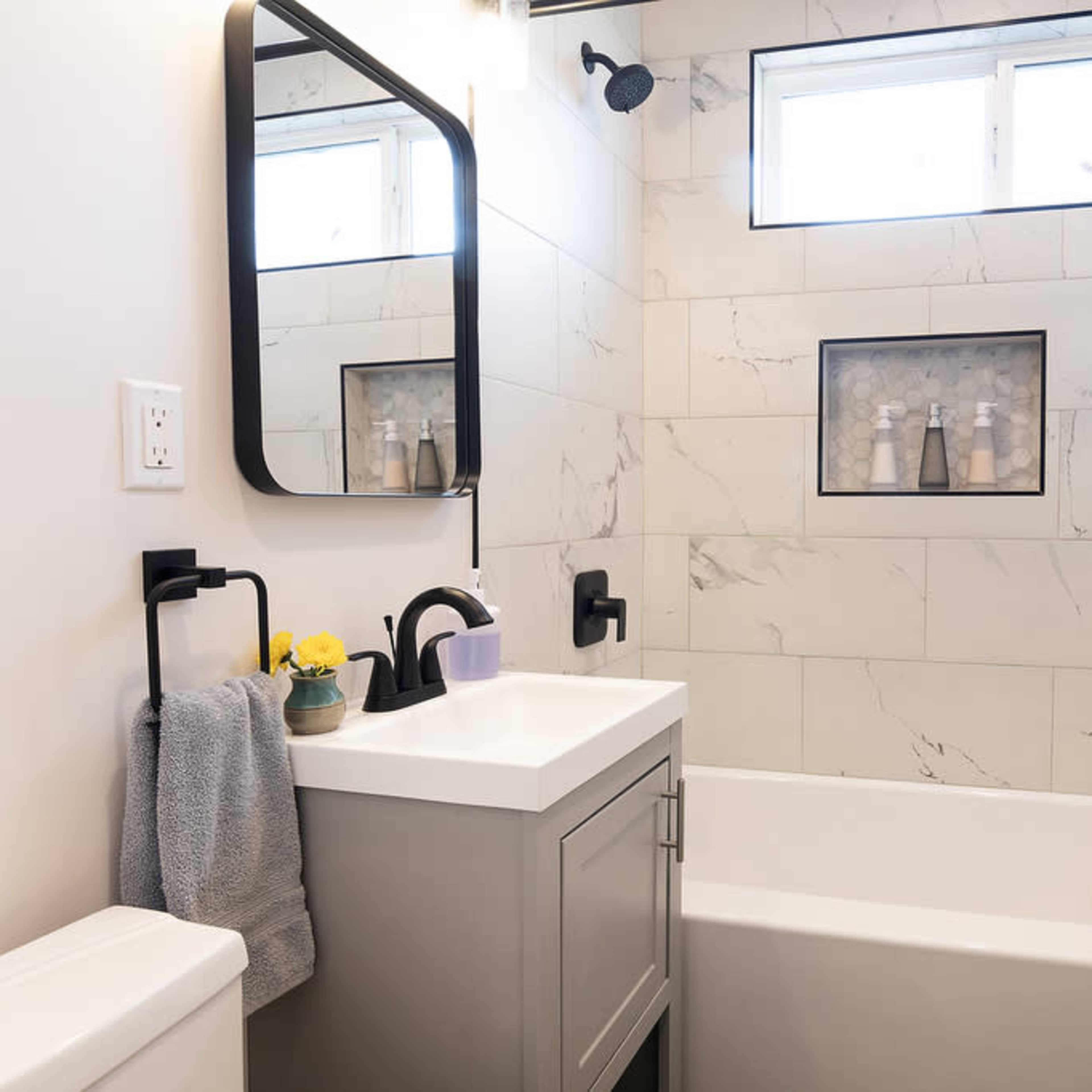 A modern bathroom features a marble-tiled shower area with a gray vanity, wall-mounted mirror, and neatly arranged toiletries.