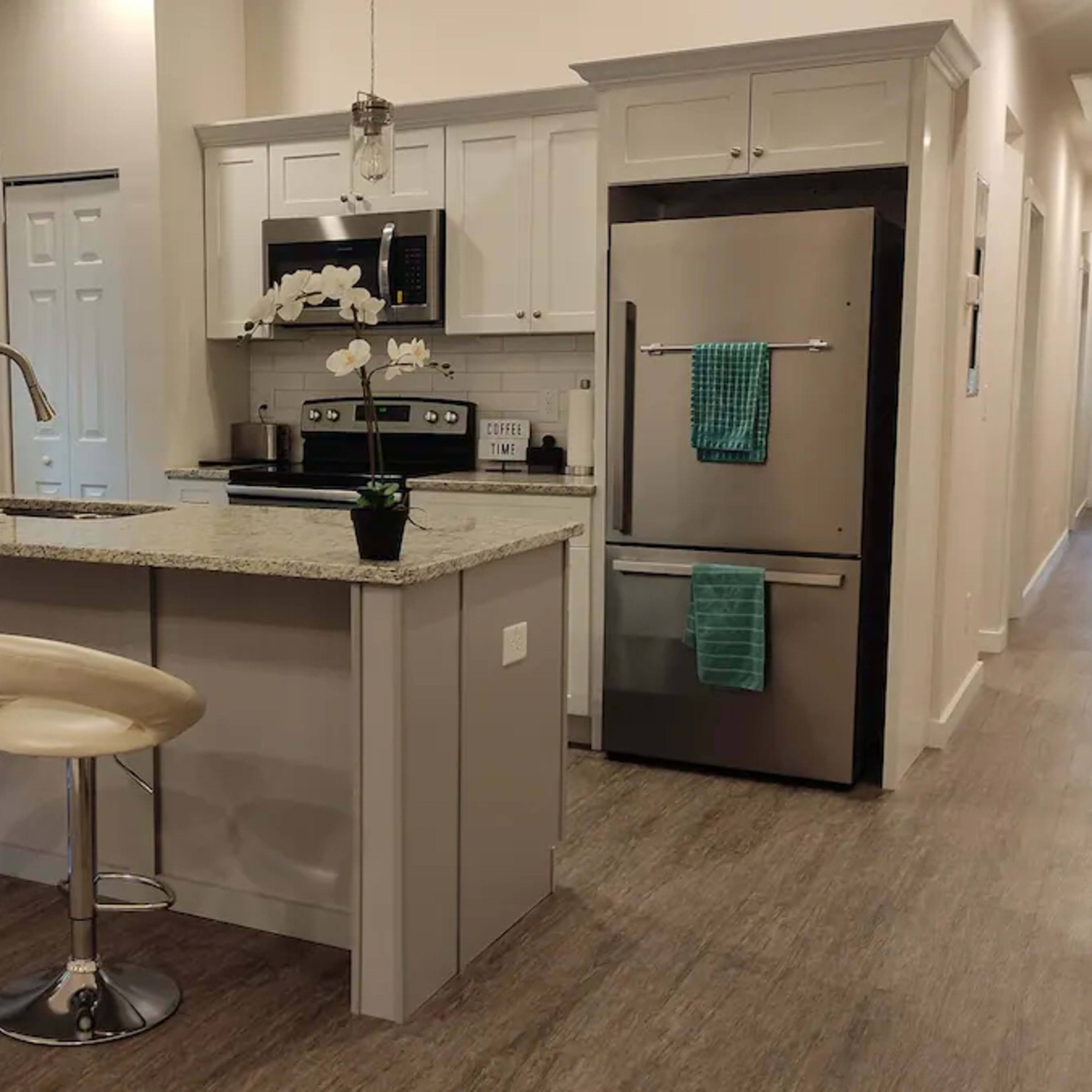 A modern kitchen with bar stools, granite countertops, stainless steel appliances, and a hallway leading to a door at the end.