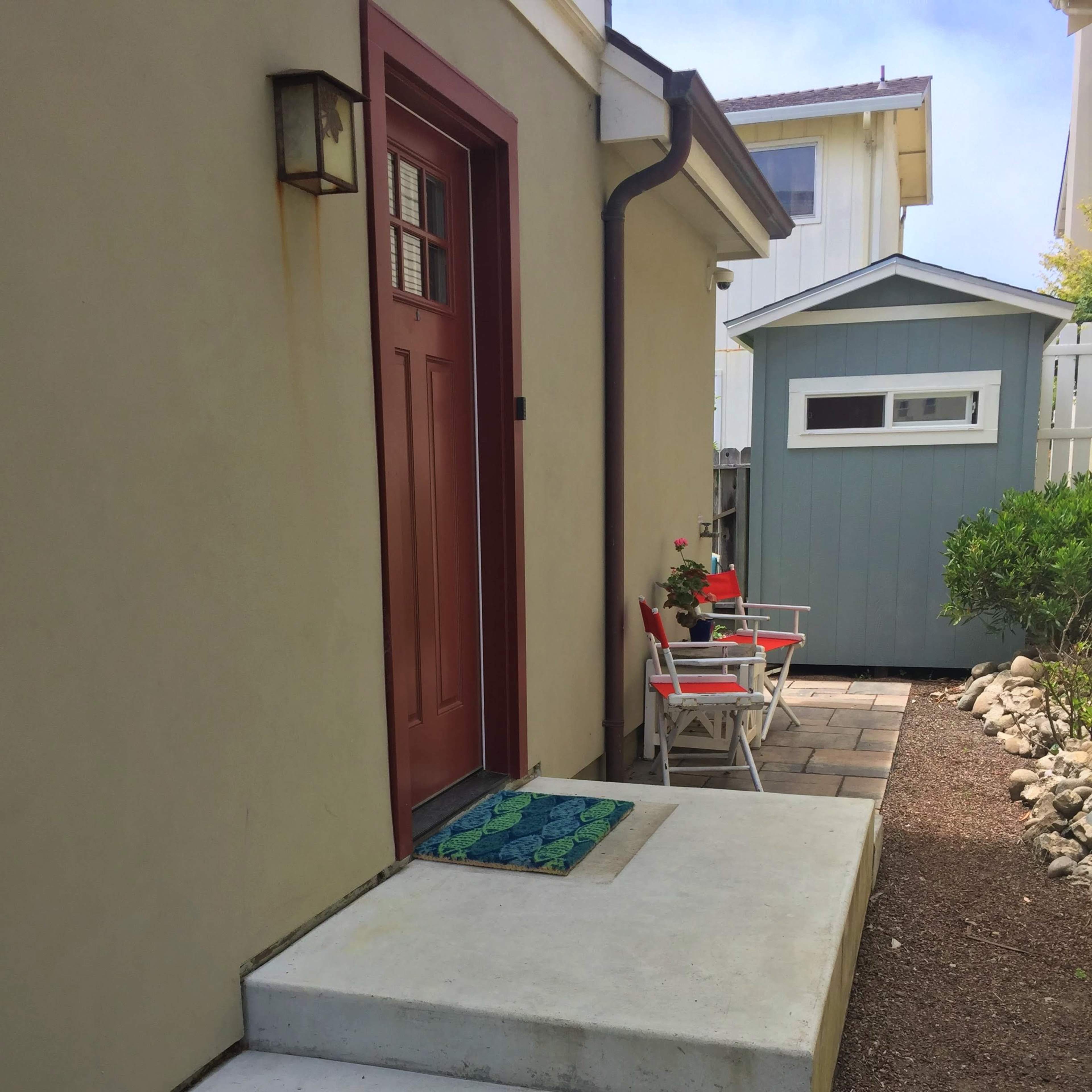A concrete entryway with a red door, flanked by a small patio area featuring two chairs and a garden shed in the background.