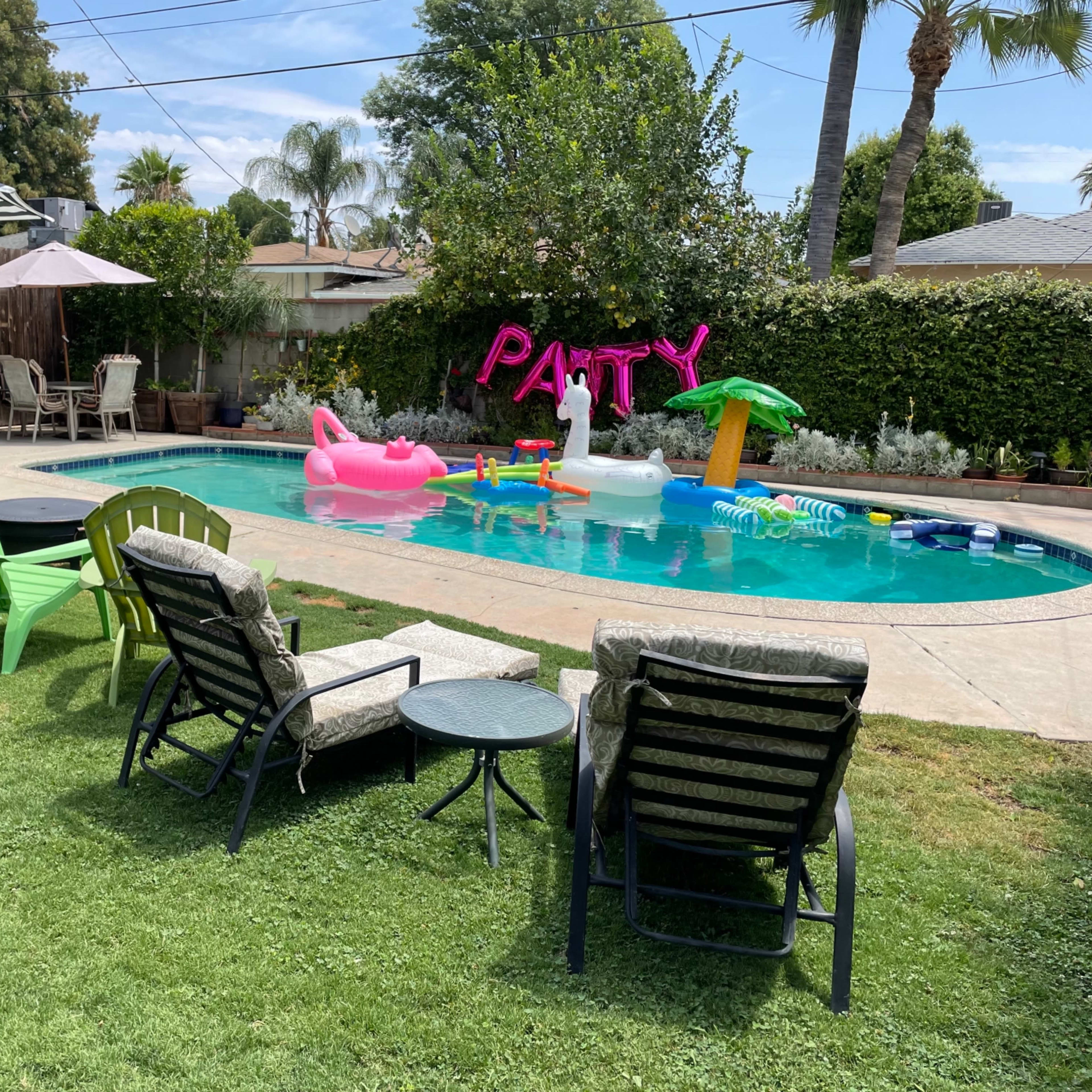 A backyard scene features a swimming pool surrounded by green lawn chairs, an umbrella, and inflatable decorations in the water.
