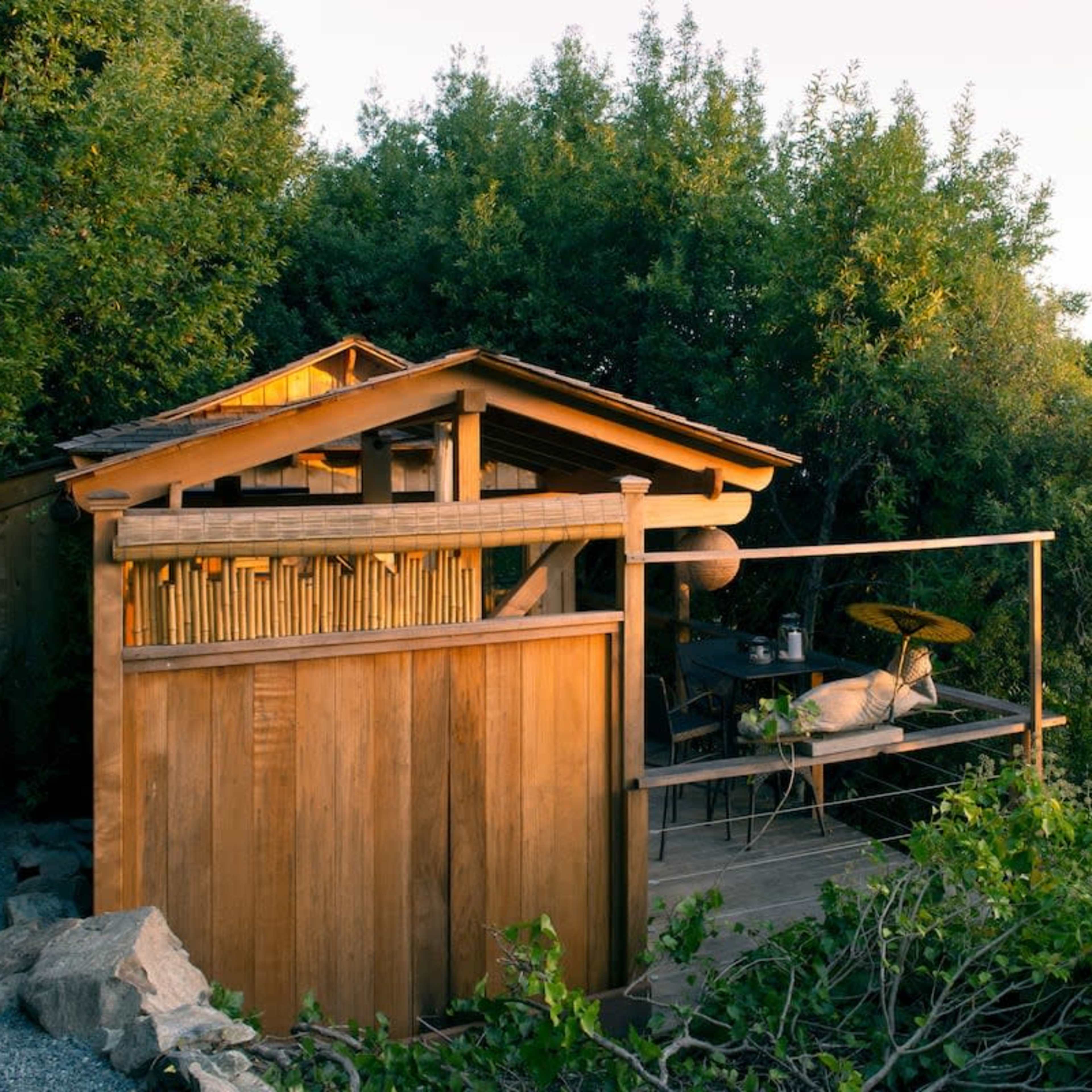 A small wooden cabin is situated among trees, with a deck overlooking a coastal landscape.