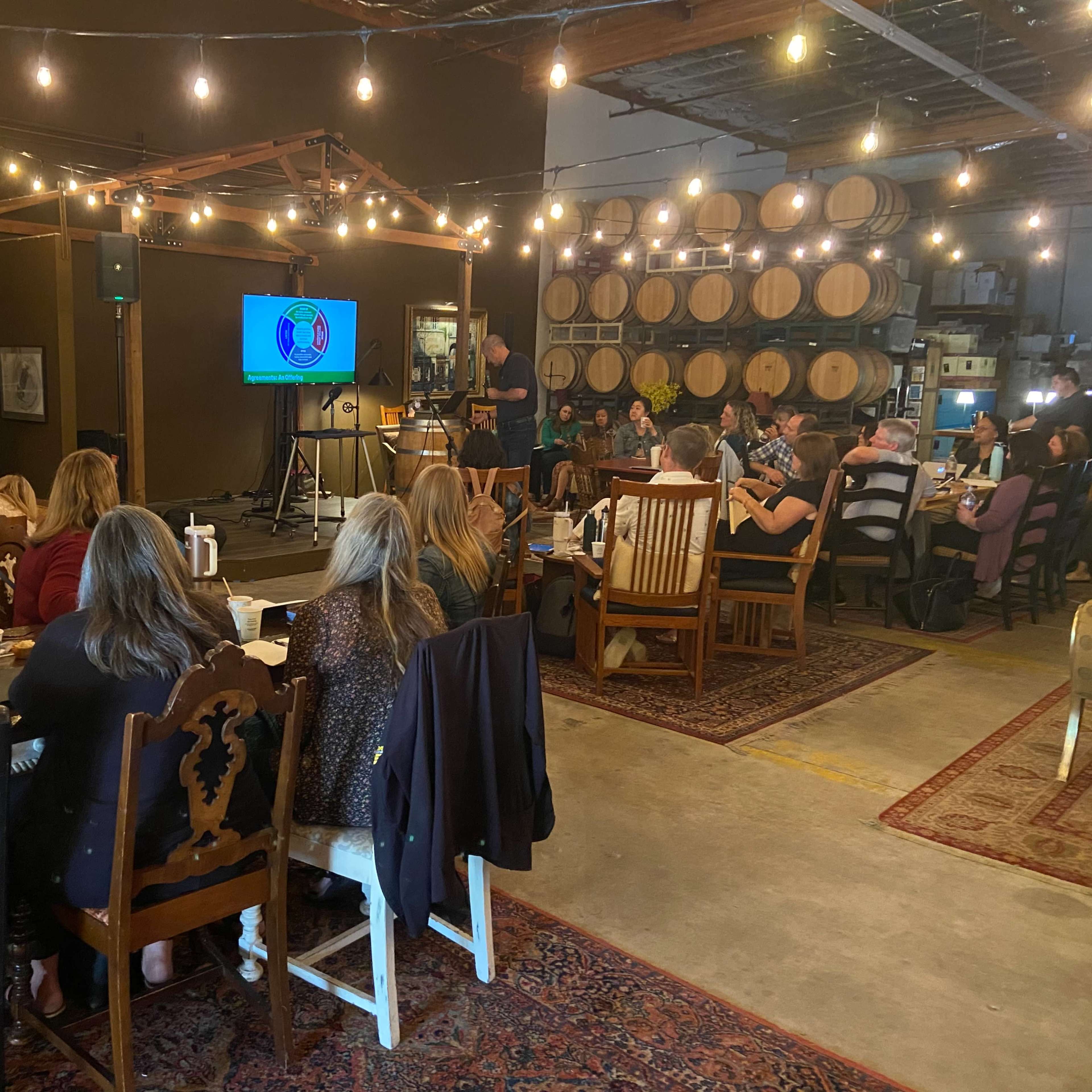 A gathering takes place inside a rustic venue with wooden barrels in the background and a presentation screen at the front.