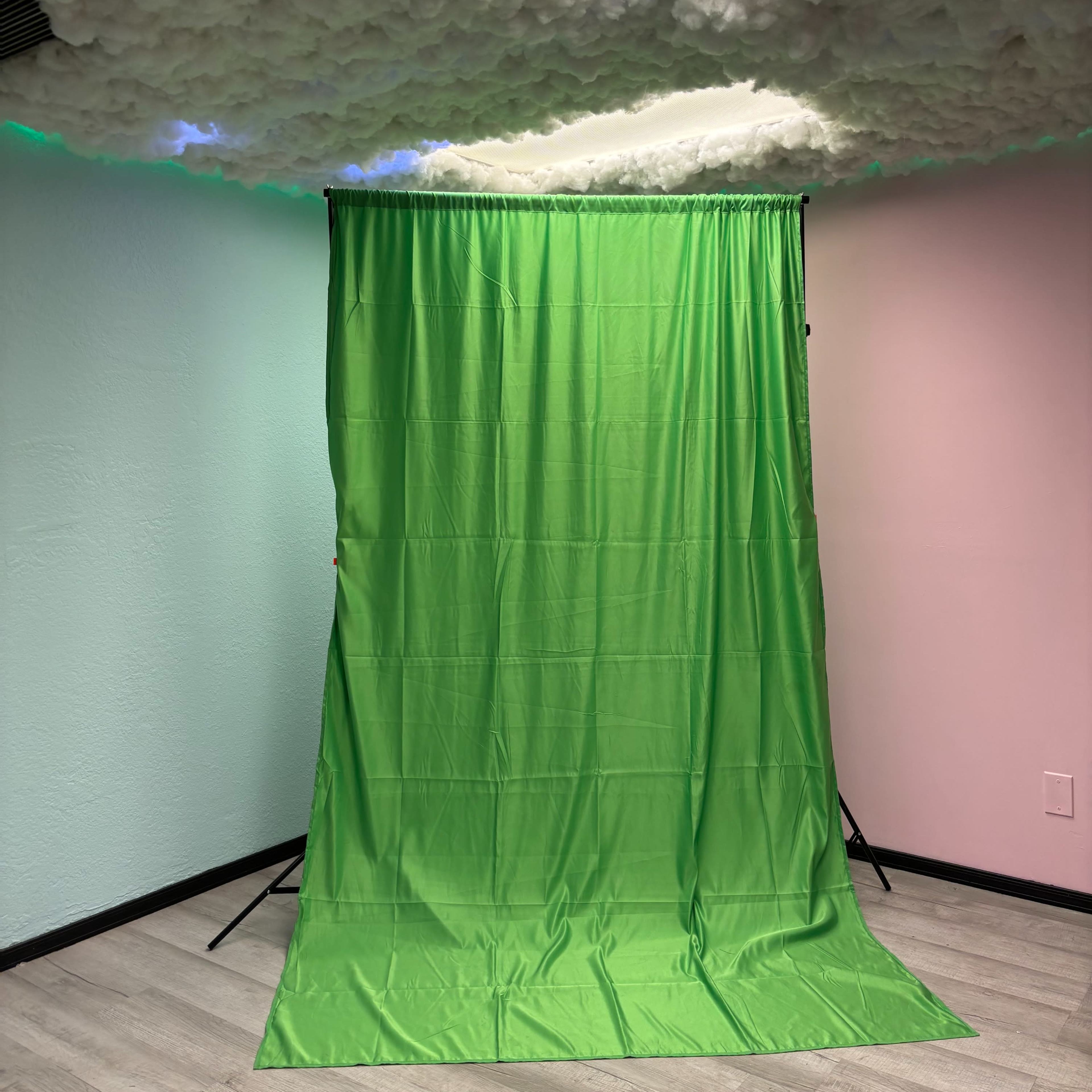 A green backdrop on a stand, set against a wall with a light pink color, under a ceiling featuring fluffy cloud-like decorations.