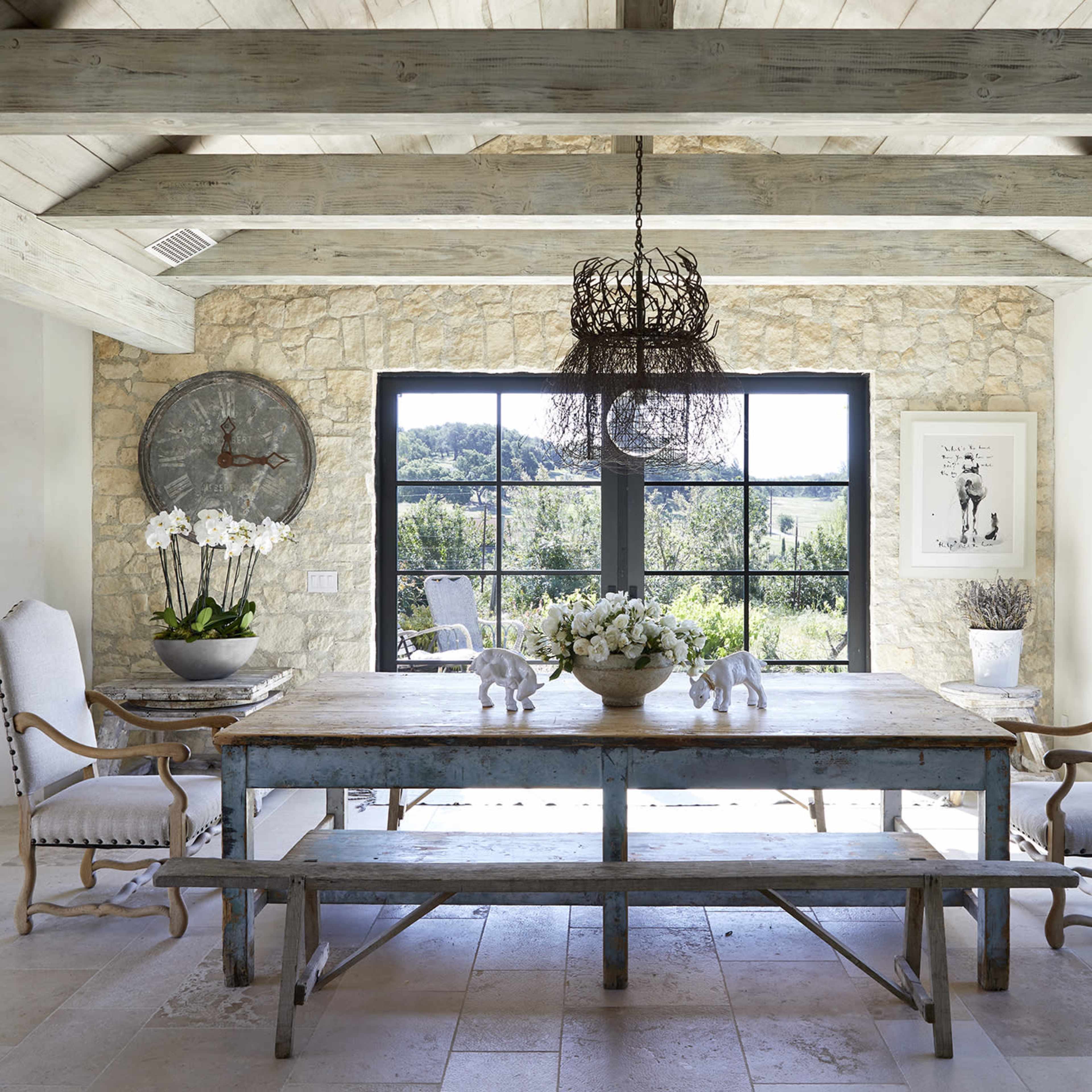 A rustic dining area featuring a wooden table, two chairs with armrests, a large clock on the wall, and large windows providing a view of the outdoors.