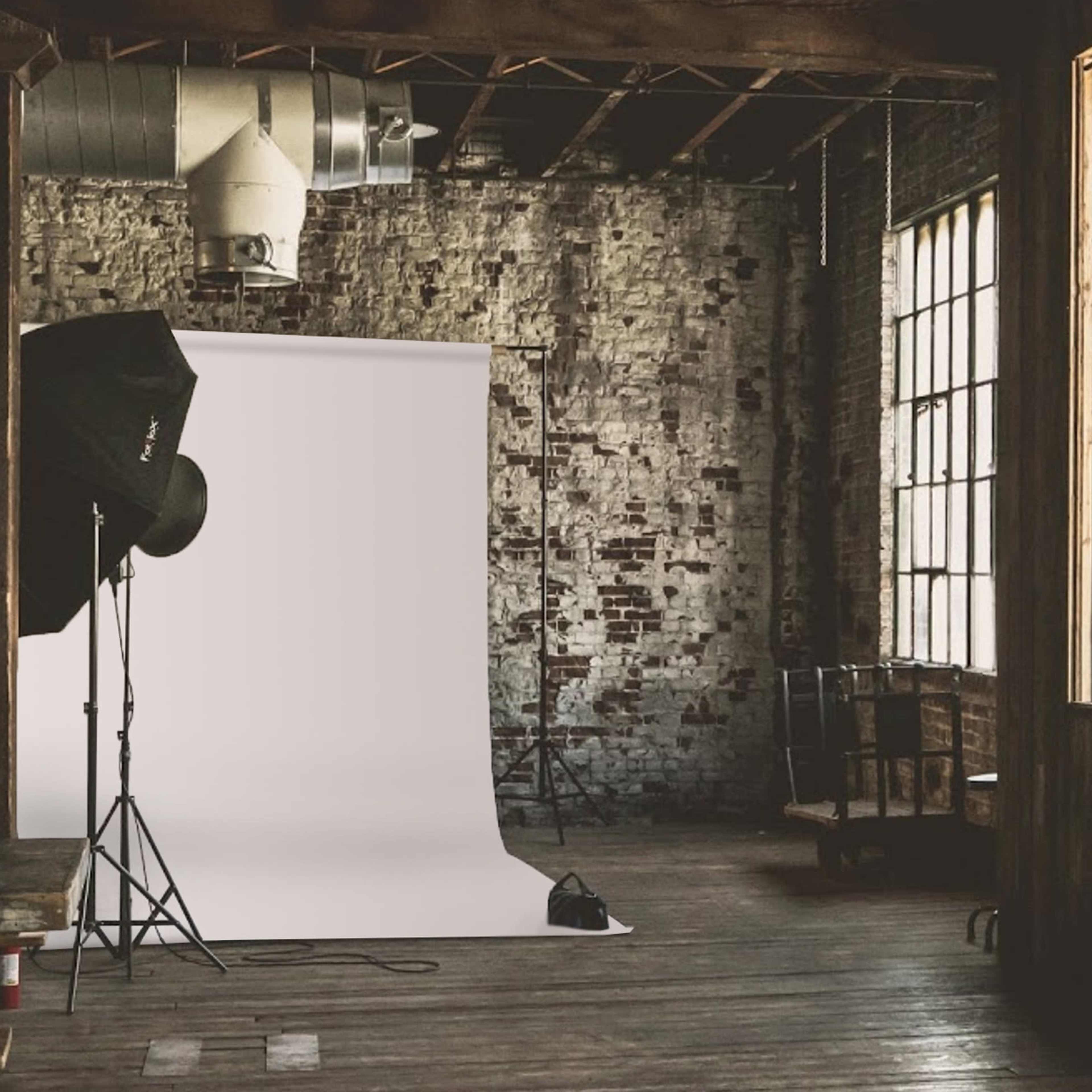 An empty industrial studio space with a white backdrop set up against a brick wall and large windows.