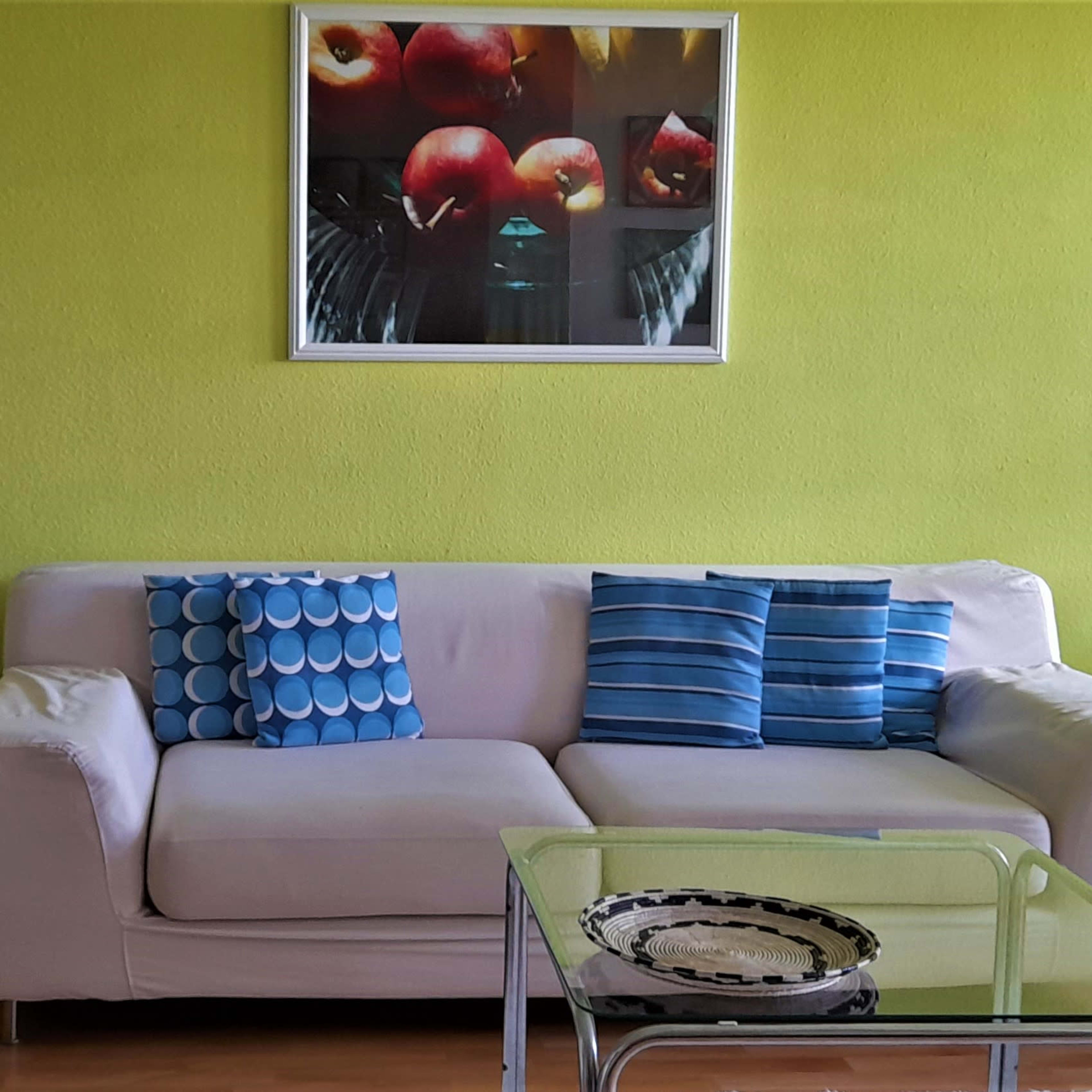 A living room features a white couch with blue and green cushions, a glass coffee table, and a vibrant green wall with a fruit-themed artwork.