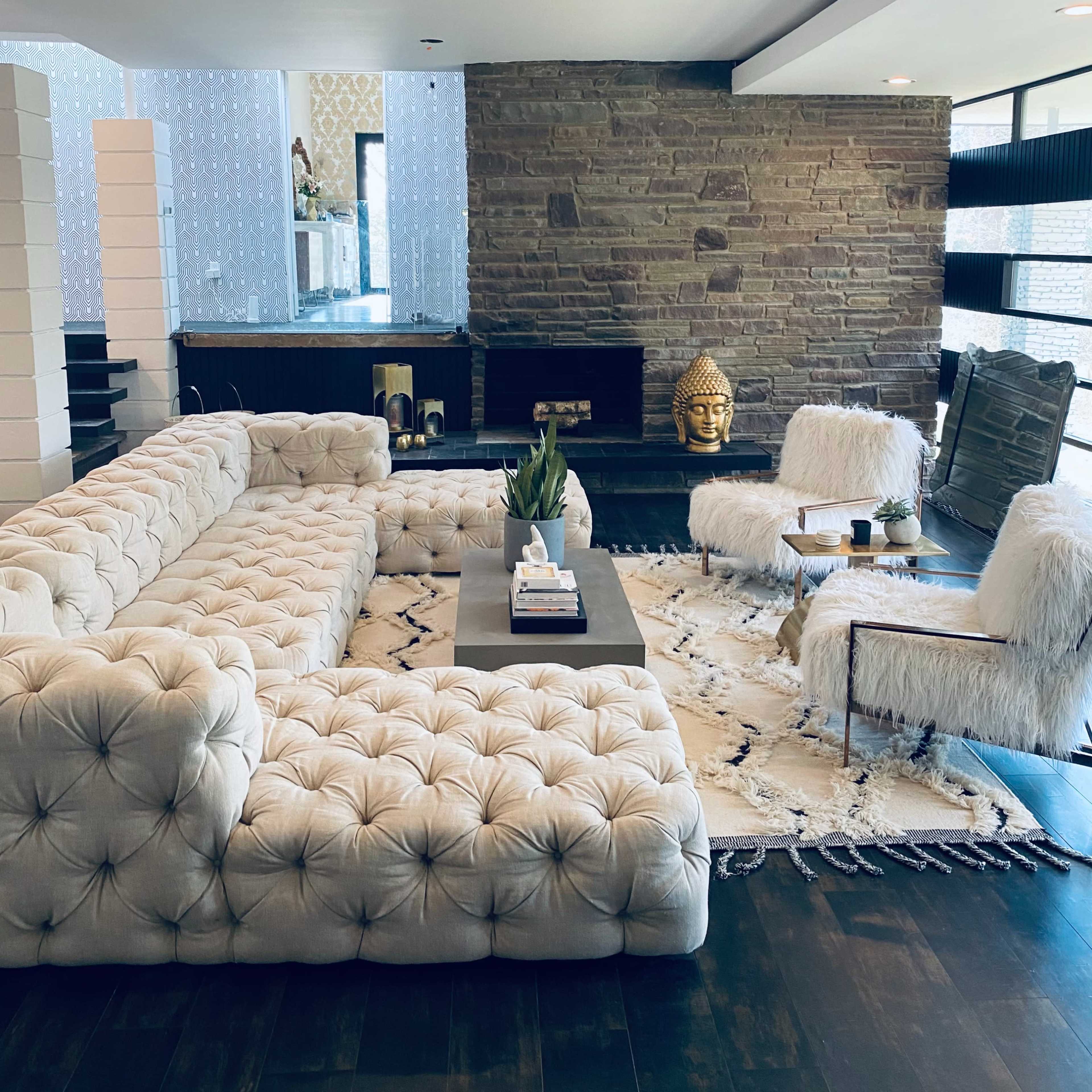A modern living room featuring a large tufted sectional sofa, two accent chairs, a stone fireplace, and a decorative area rug.