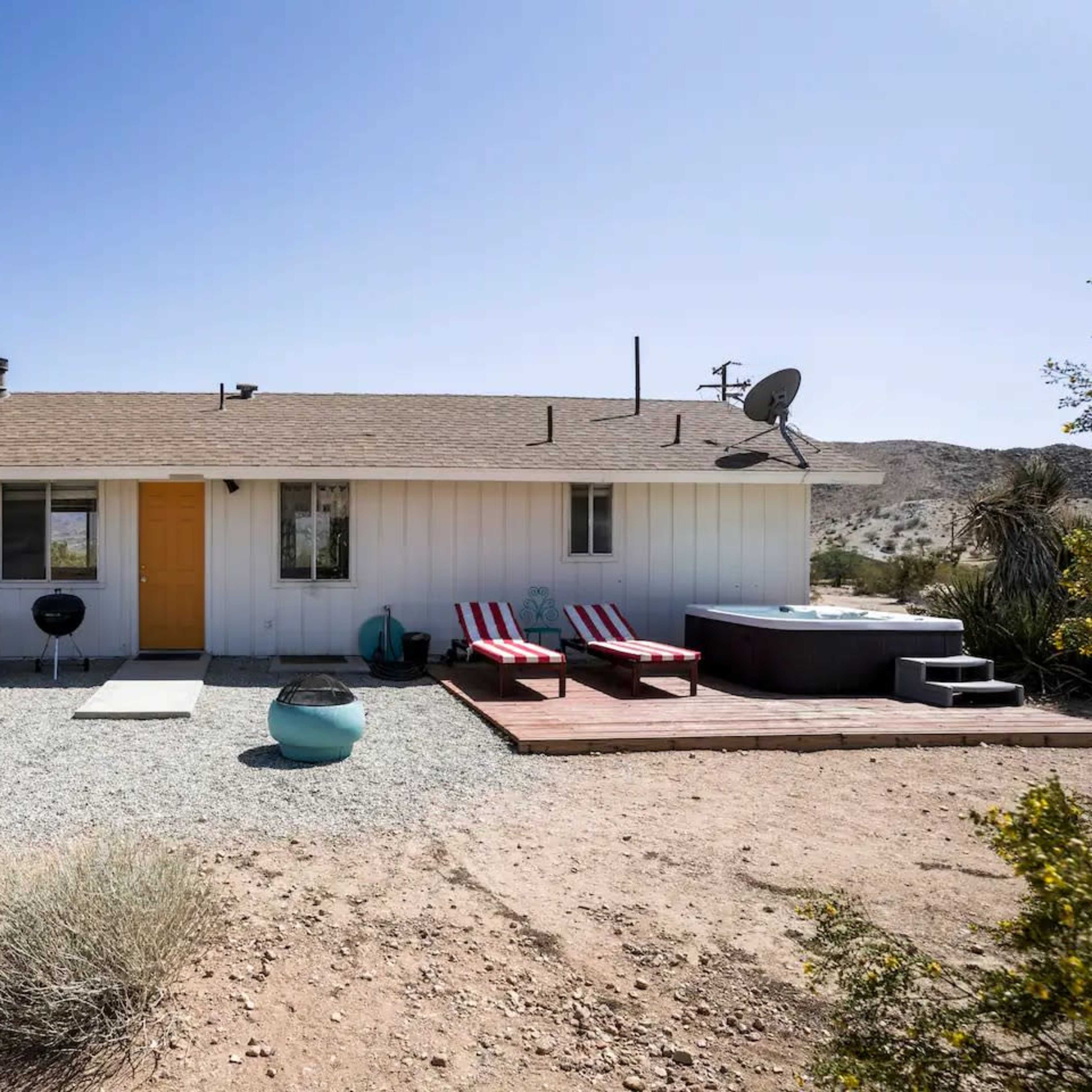 A single-story white house with orange trim and a wooden deck is surrounded by desert landscaping and outdoor furniture.