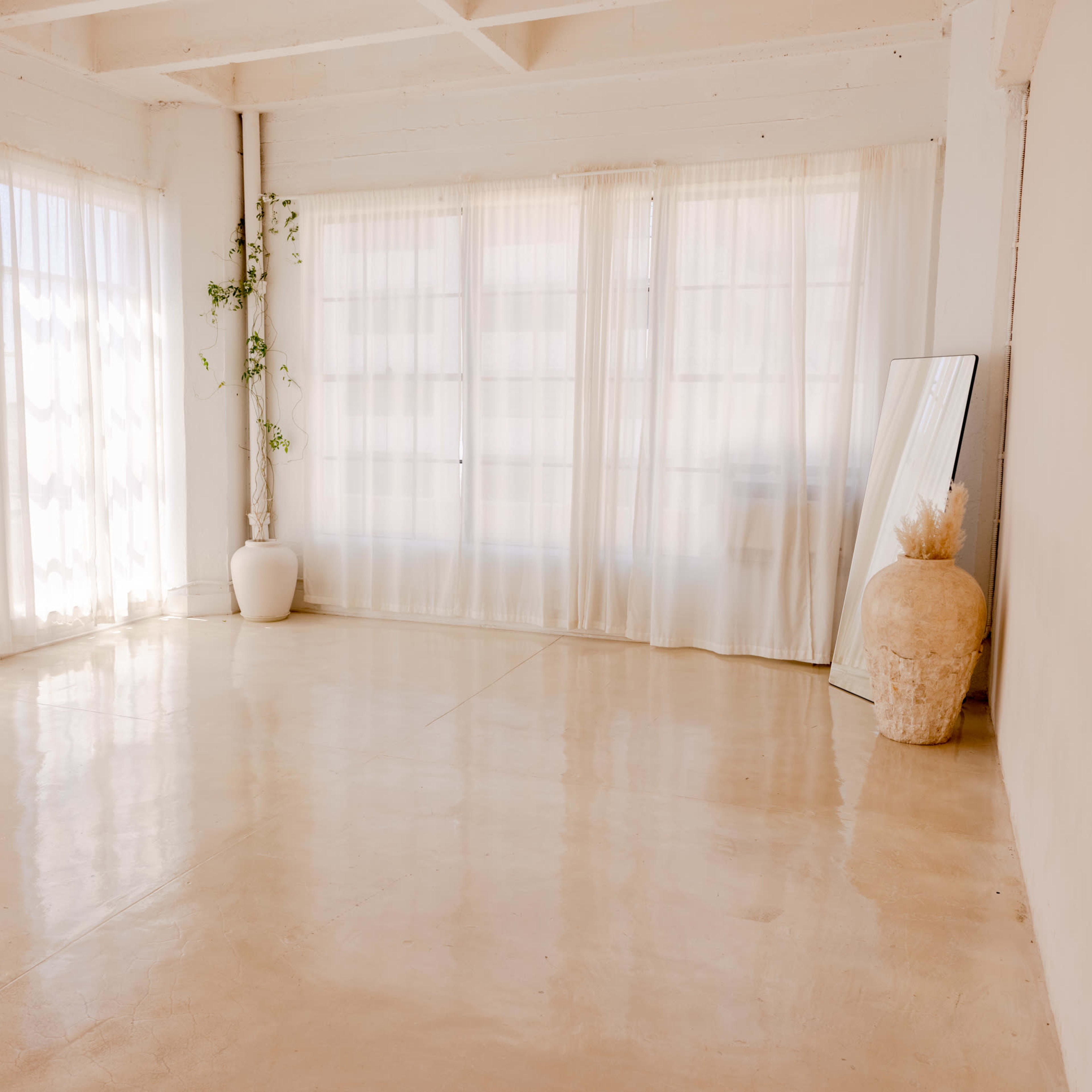 A bright, empty room with large windows covered by sheer curtains and a few decorative items, including a potted plant and a mirror.