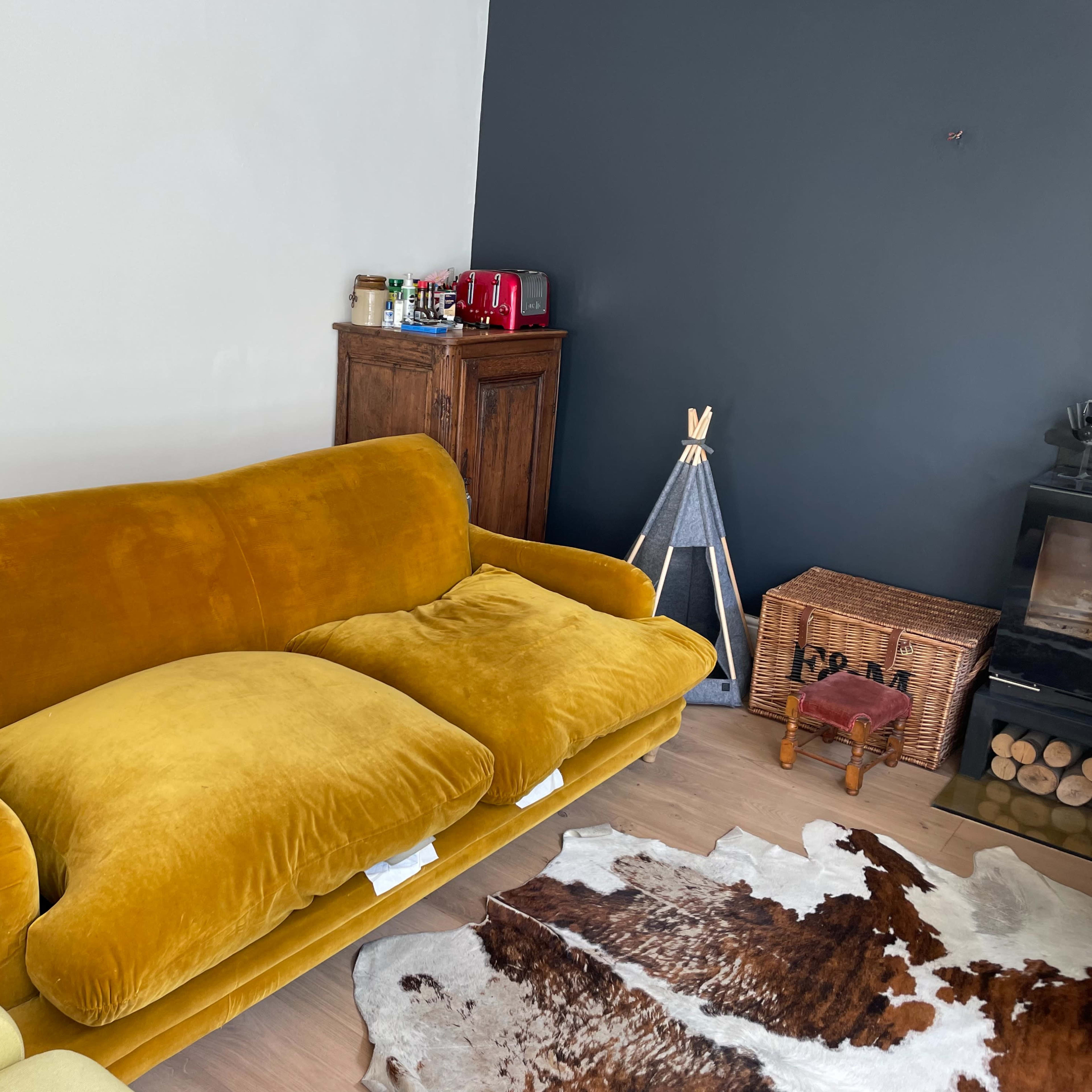 A yellow velvet couch with two oversized cushions sits against a white and dark gray wall, accompanied by a wooden cabinet and a small decorative structure in the corner.