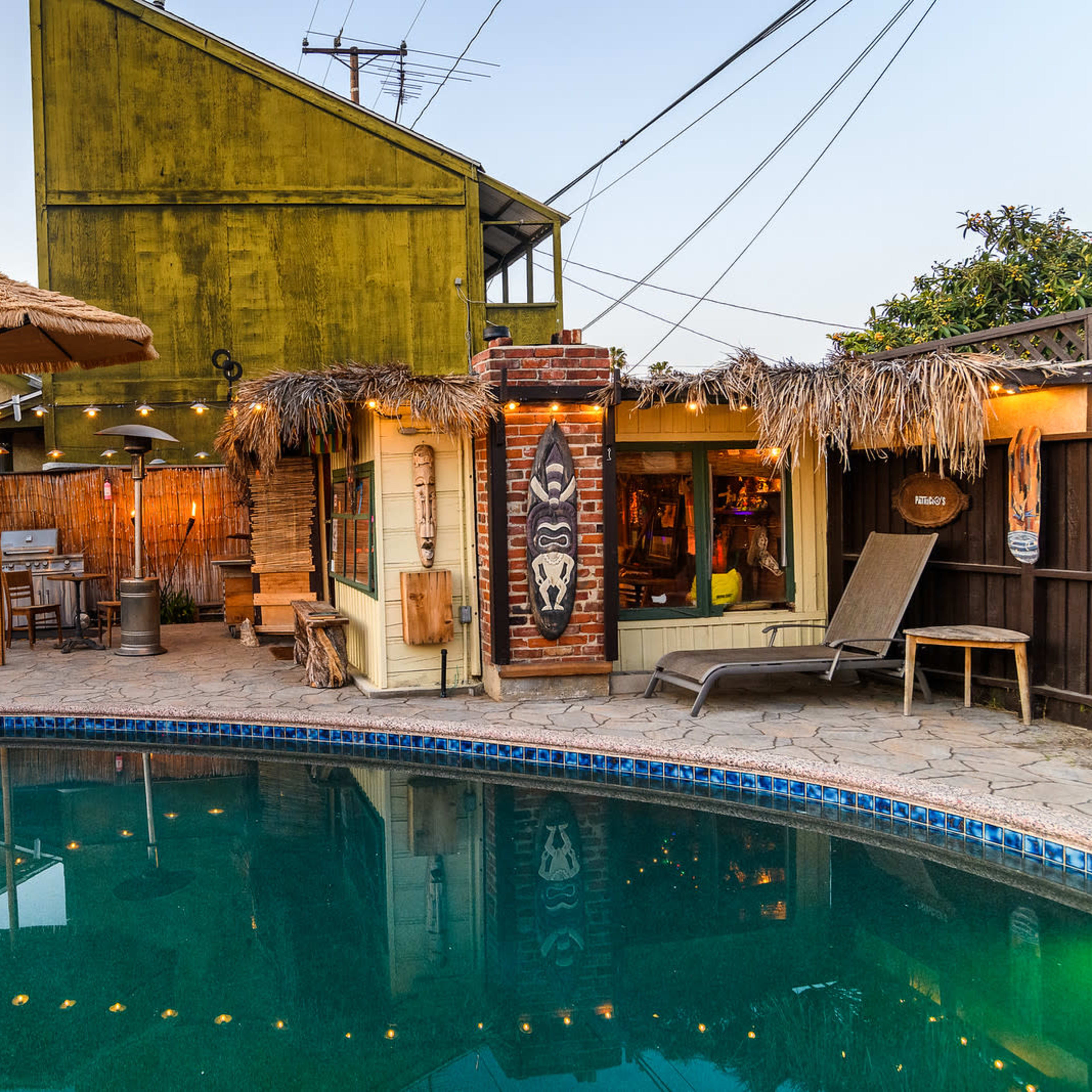 A backyard pool area featuring a green house, a thatched umbrella, and outdoor seating surrounded by trees and overhead power lines.