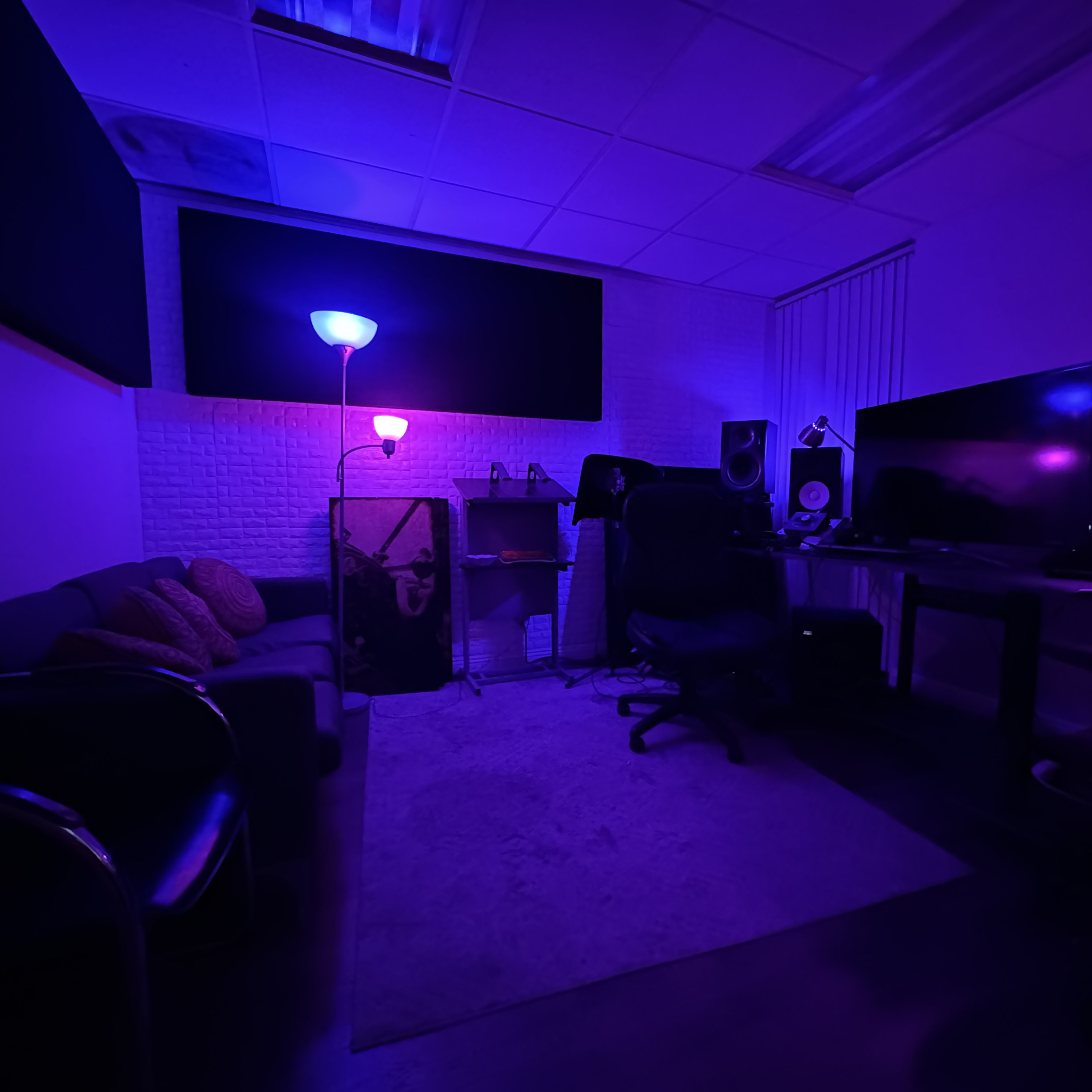 A dimly lit room features a couch, a desk with monitors, and colorful lighting from lamps, creating a workspace atmosphere.