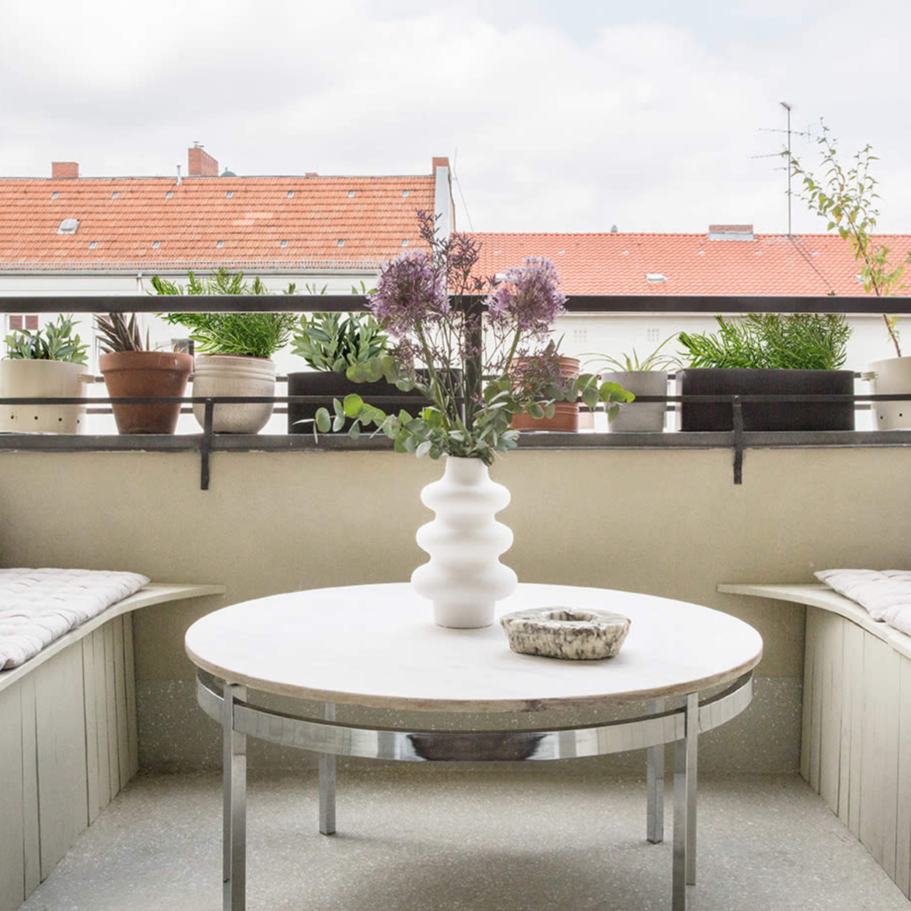 A balcony features two seating areas with cushions, a circular table in the center, and various potted plants along the railing.