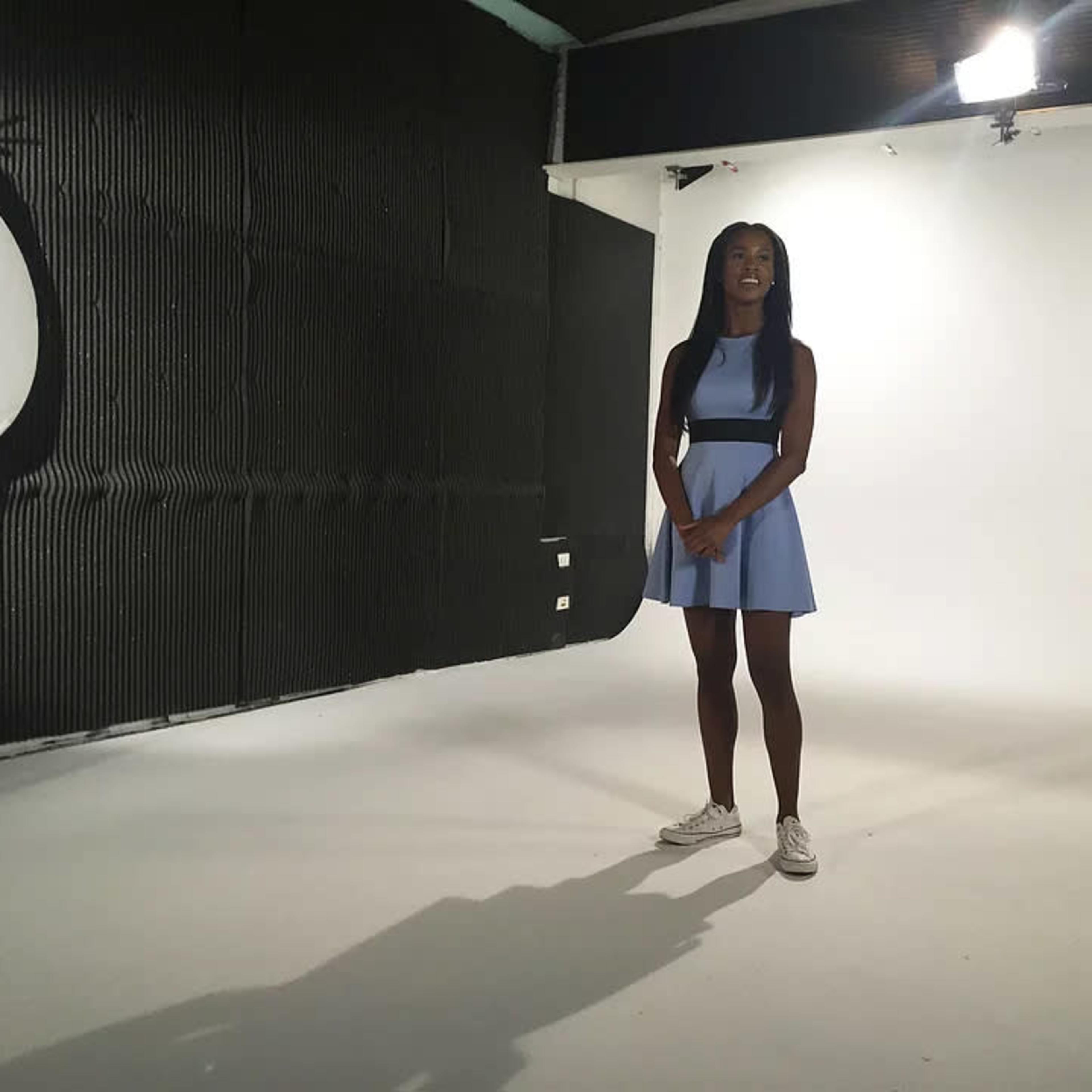 A woman stands in a well-lit photography studio, wearing a light blue dress and sneakers.