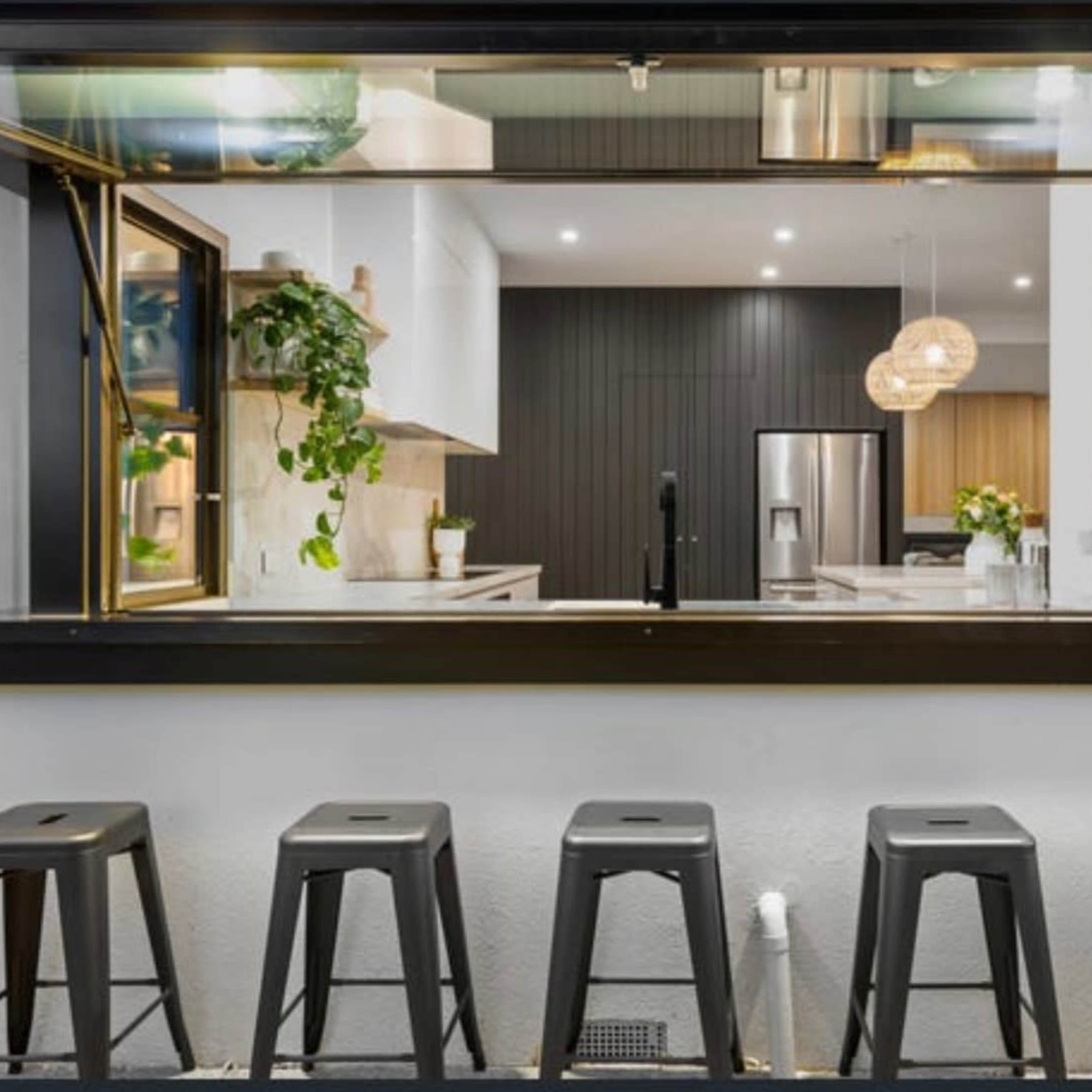 A kitchen with a large open window overlooking a row of black metal stools placed outside.