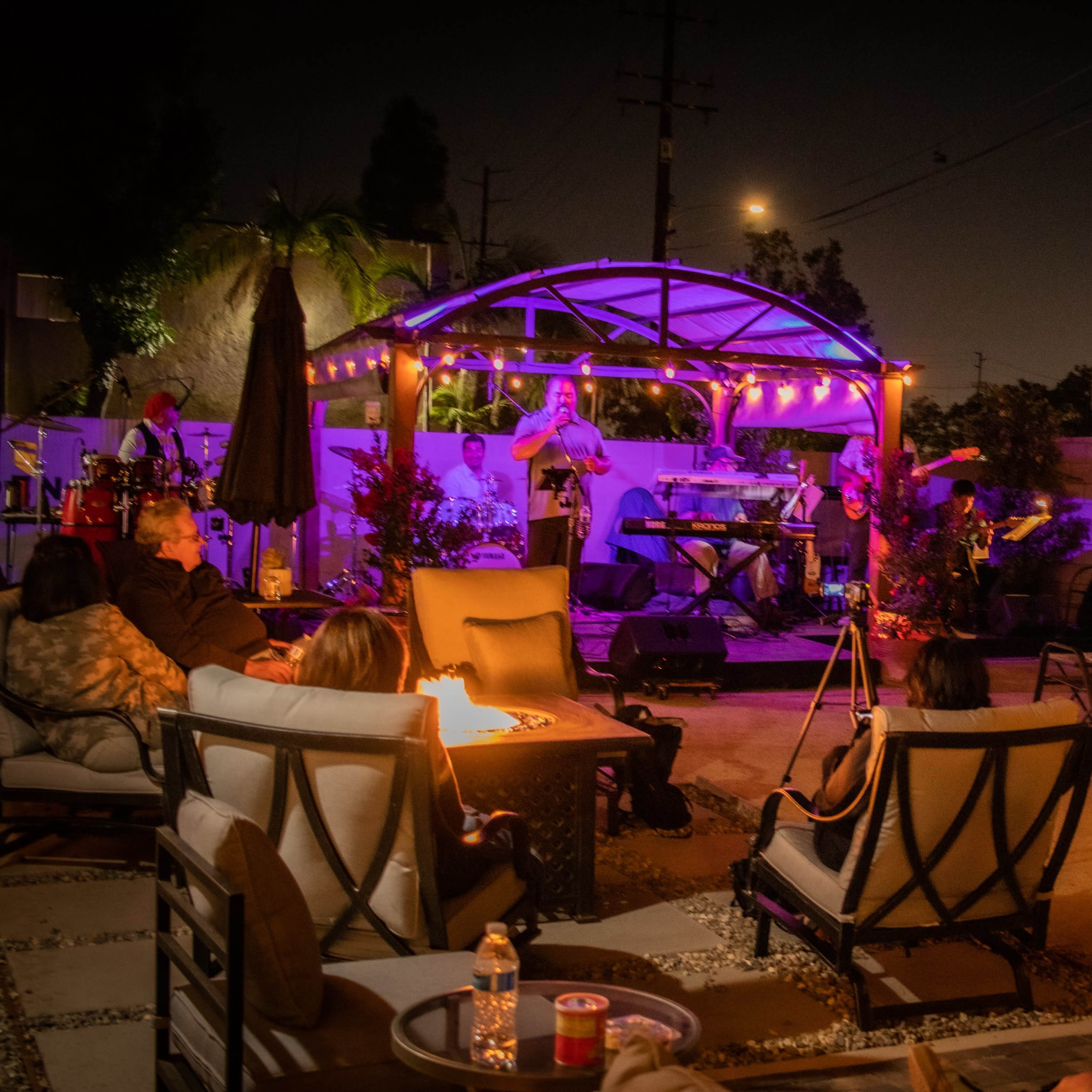 A live band performs under a lit gazebo in a backyard while guests relax around fire pits.