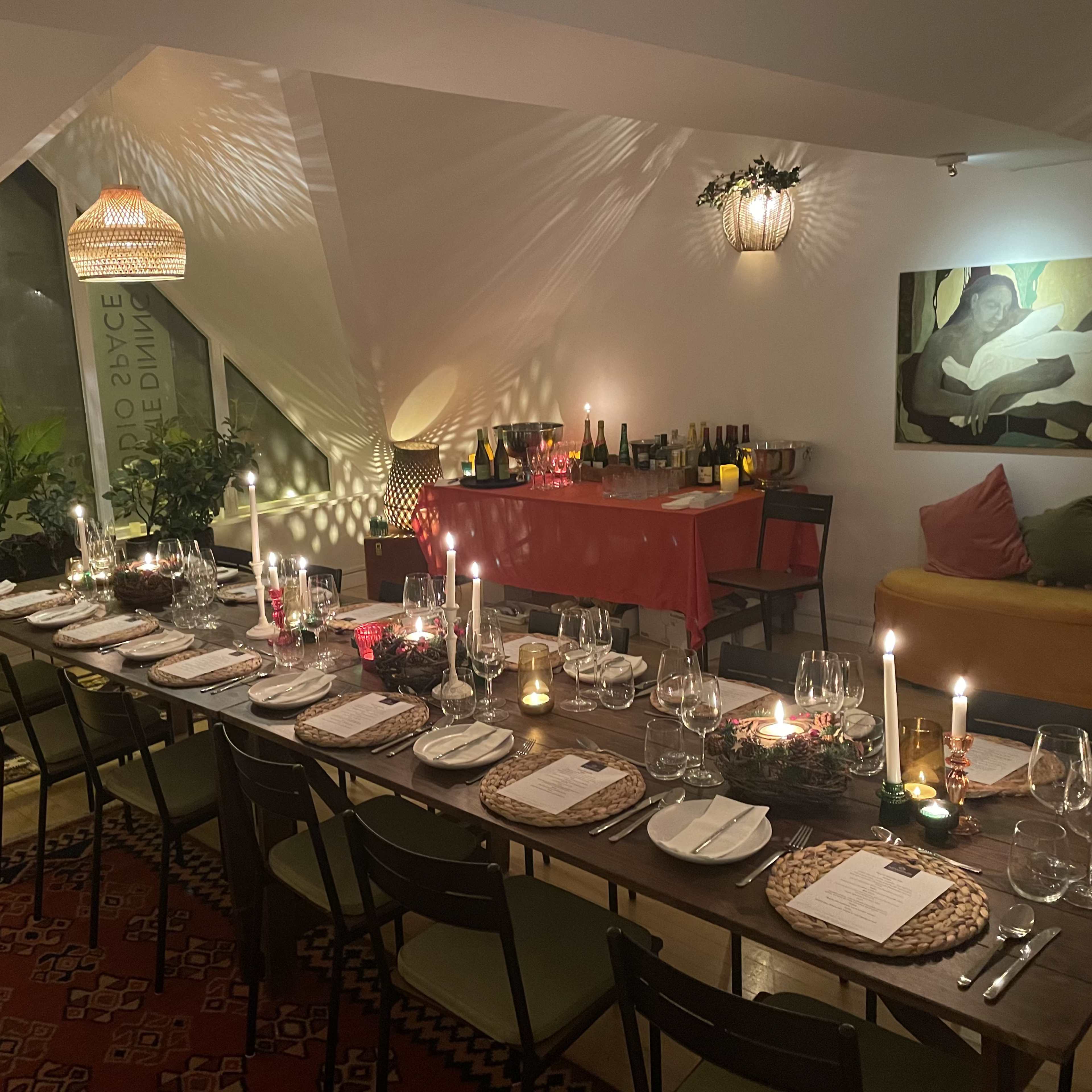 A long dining table is set with plates, glasses, and candles, surrounded by plants and a bar area with bottles in a well-lit, modern room.