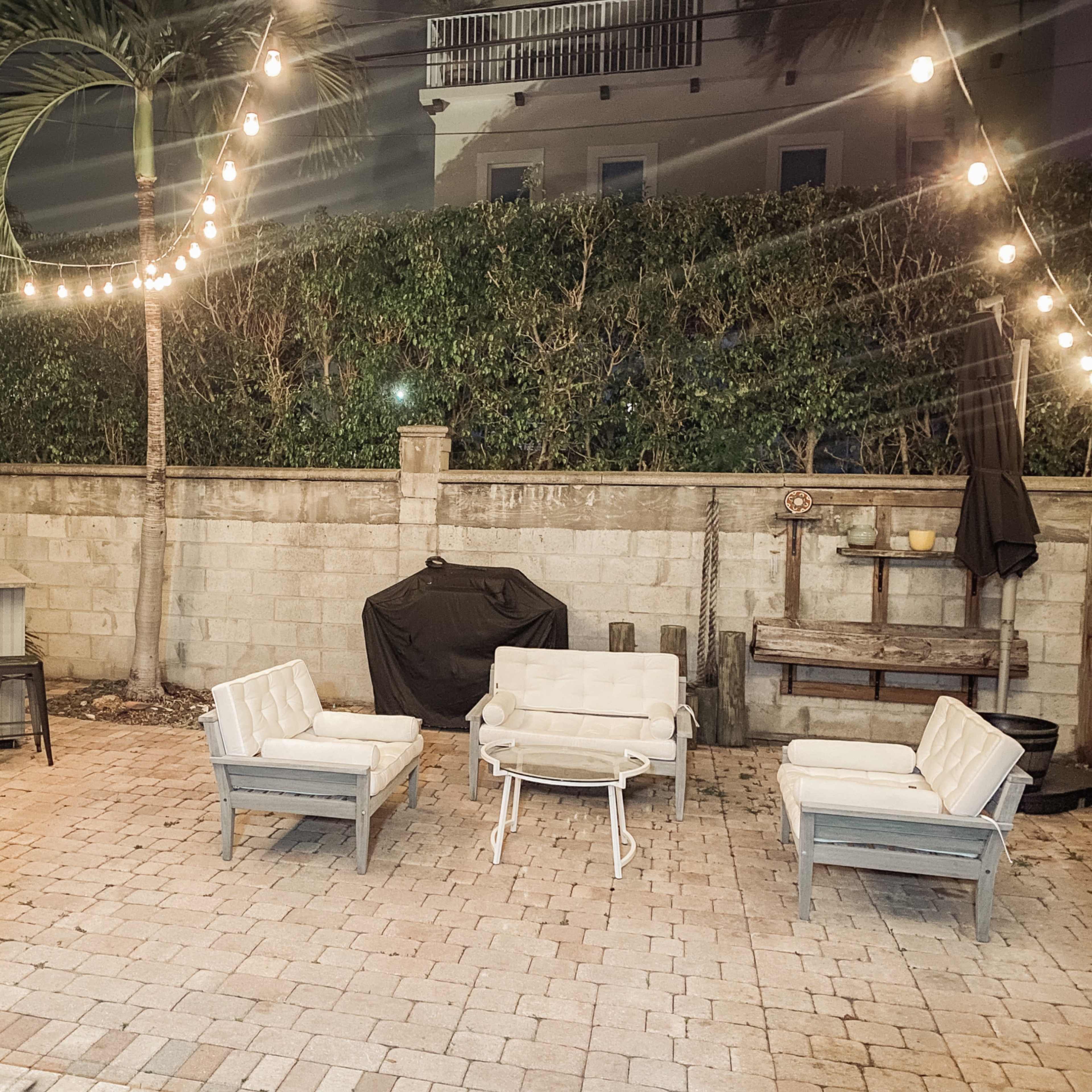 A patio area with four white chairs arranged around a small table, a bar counter with stools in the background, and string lights overhead.