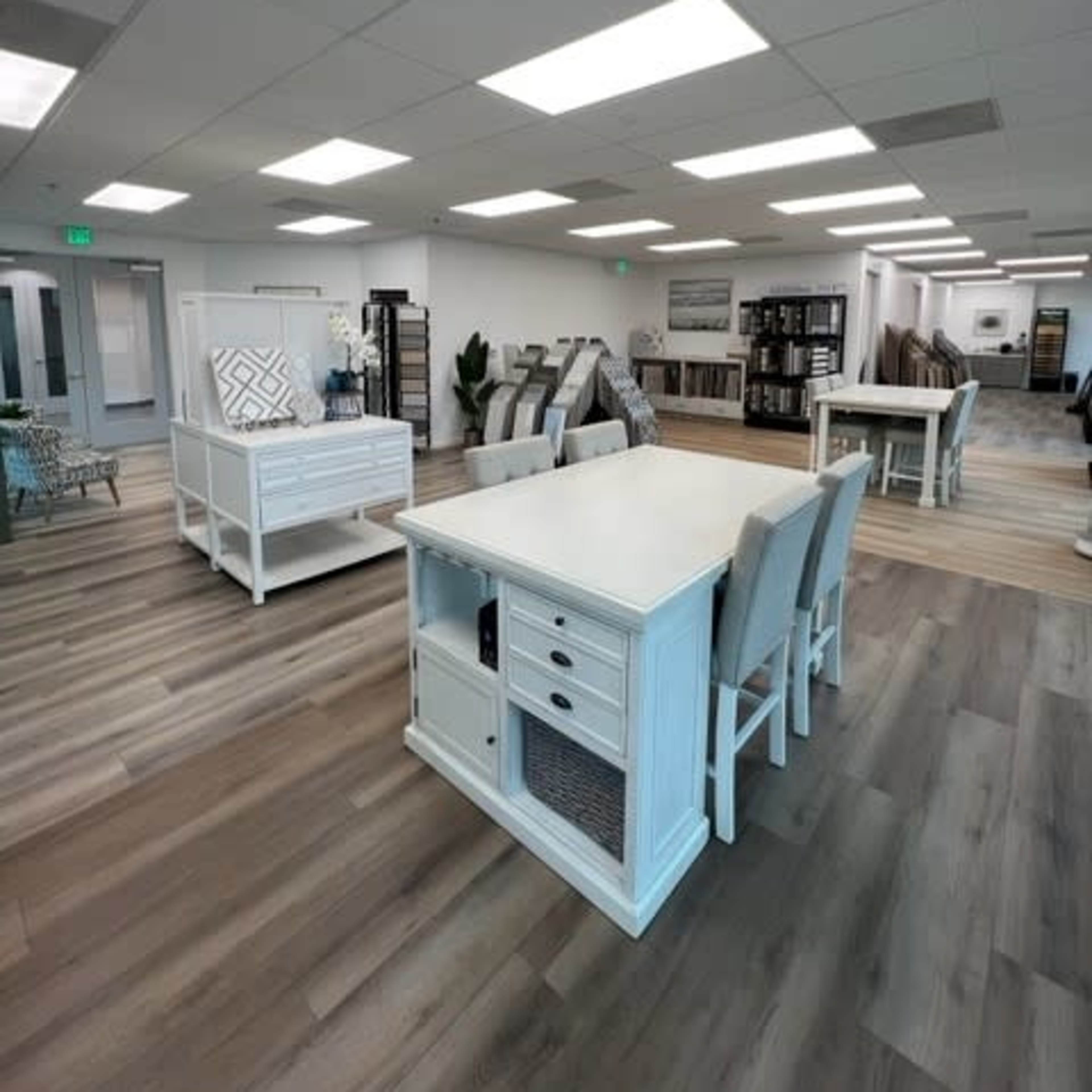 A spacious interior with white furniture, including tables and chairs, arranged in a well-lit showroom featuring wood flooring.