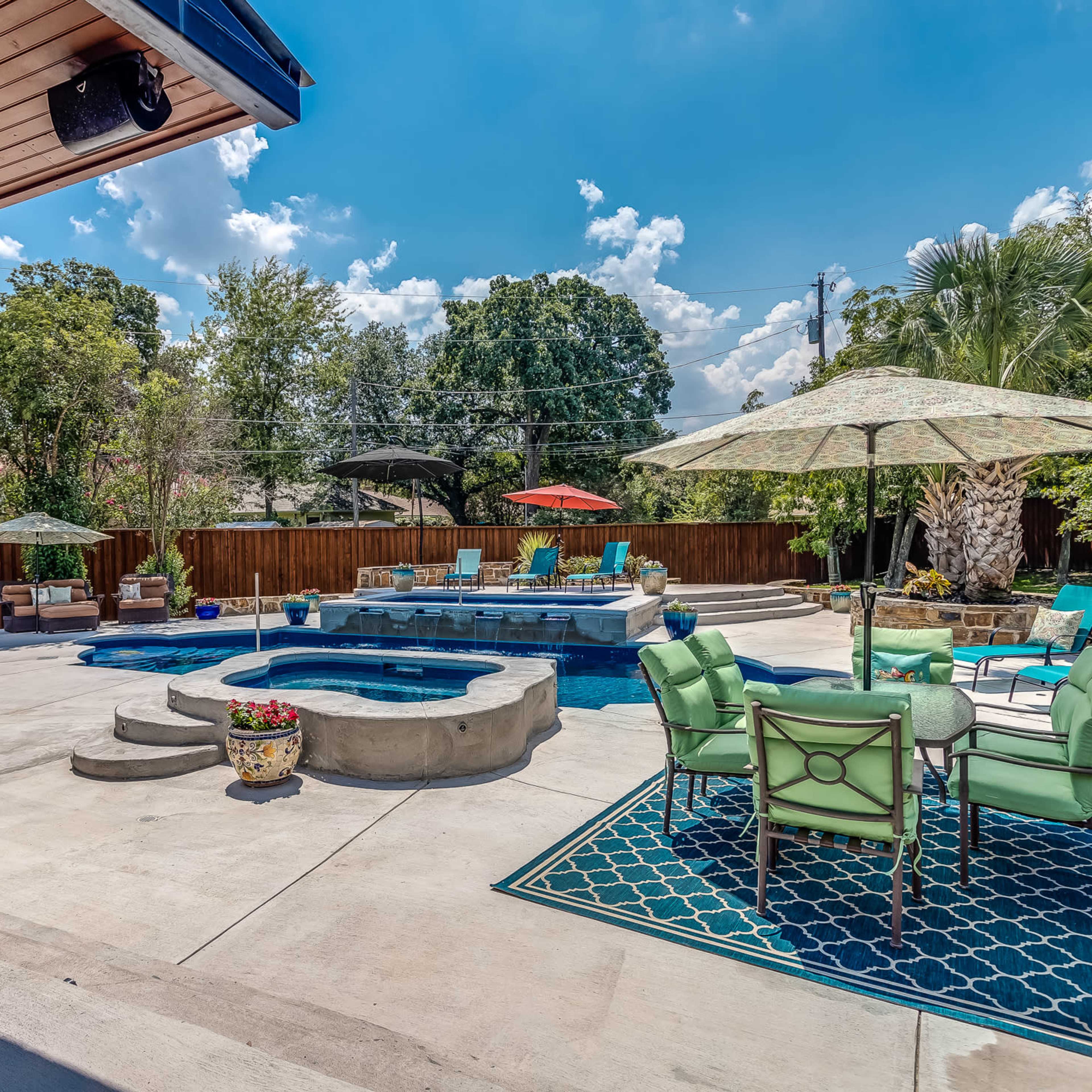 A backyard features a pool with a spa, surrounded by patio chairs, umbrellas, and landscaped greenery.
