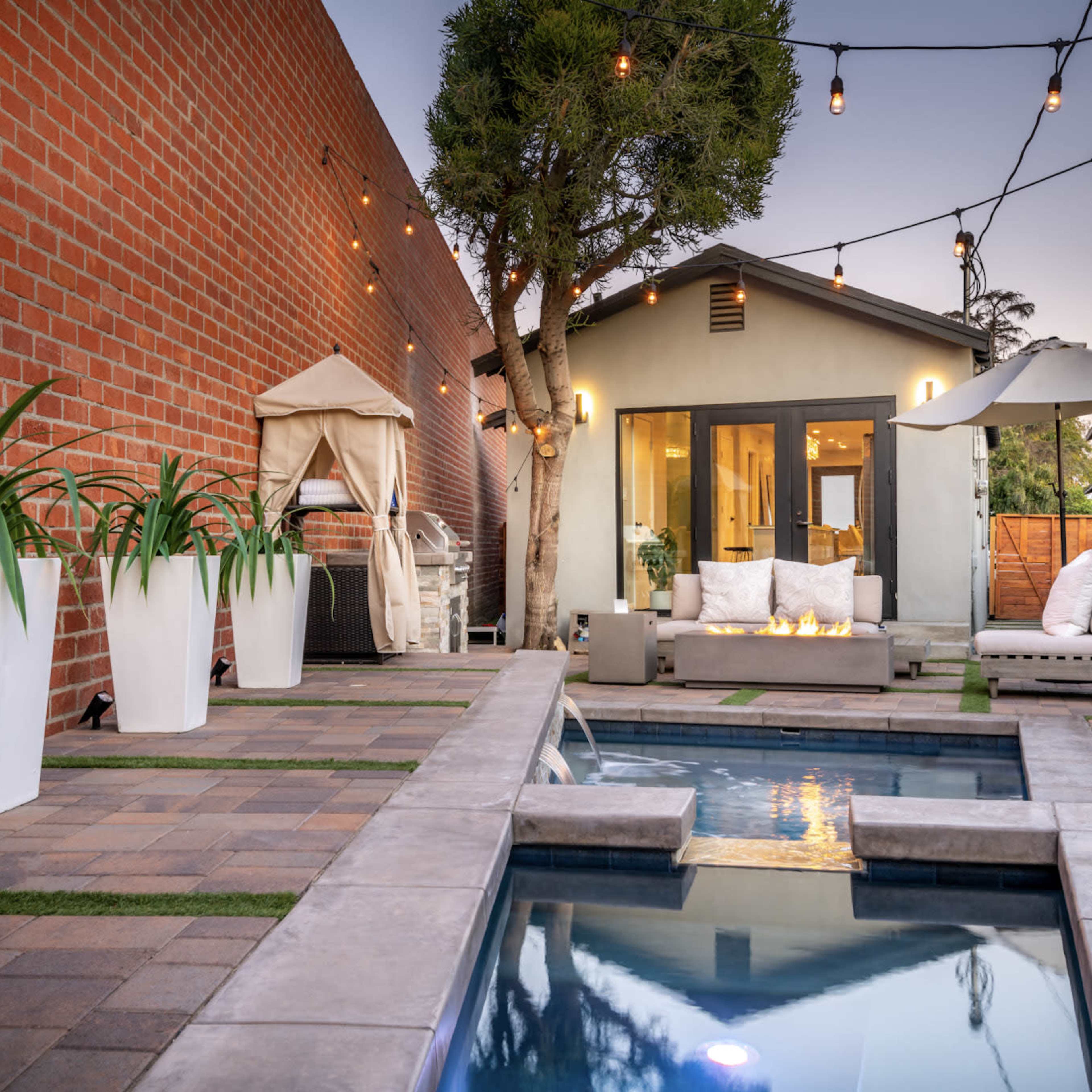 A modern outdoor patio featuring a rectangular pool, a seating area with a fire pit, large potted plants, and string lights overhead.