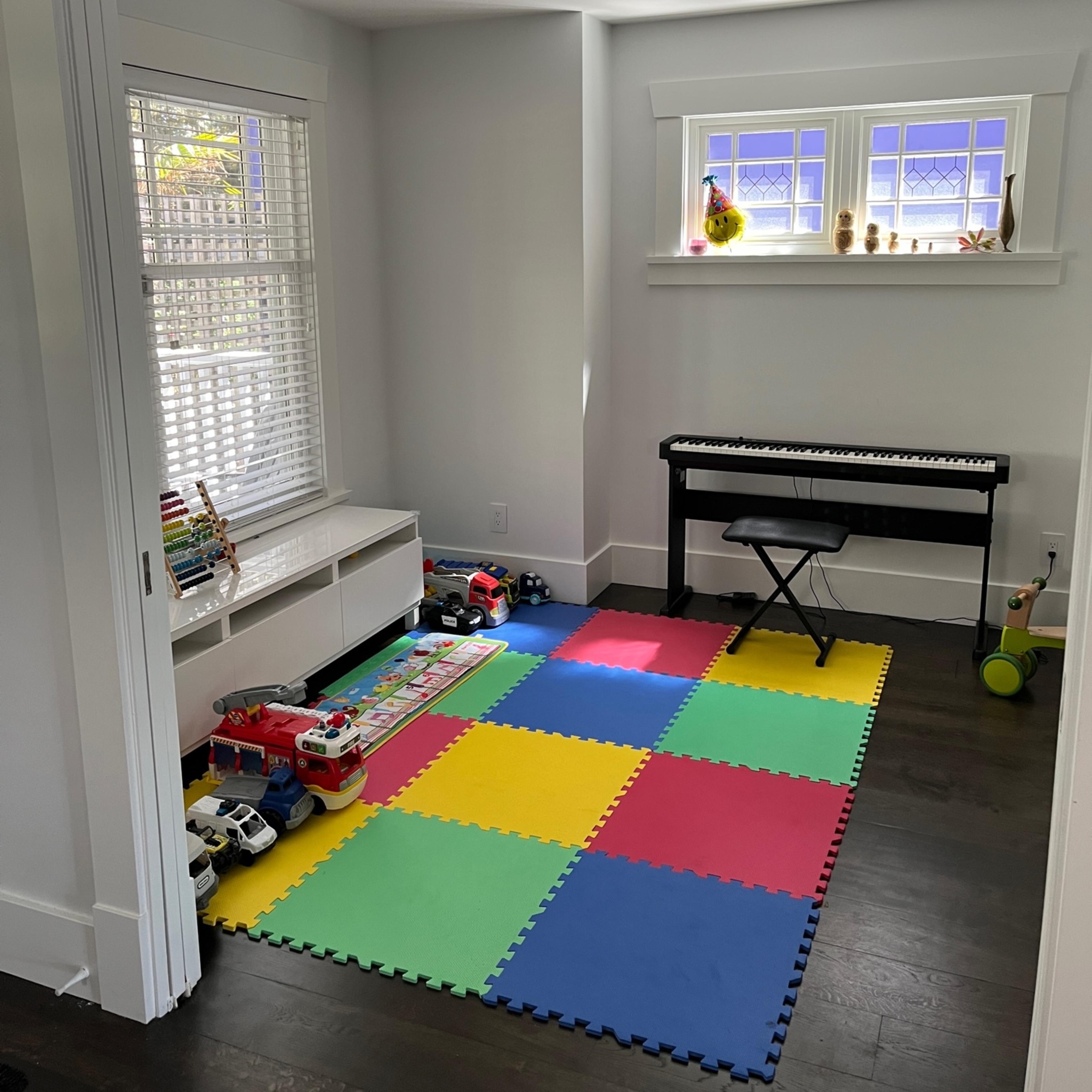 A brightly colored playroom features a patterned foam mat, a toy assortment, and a keyboard positioned beside a window.