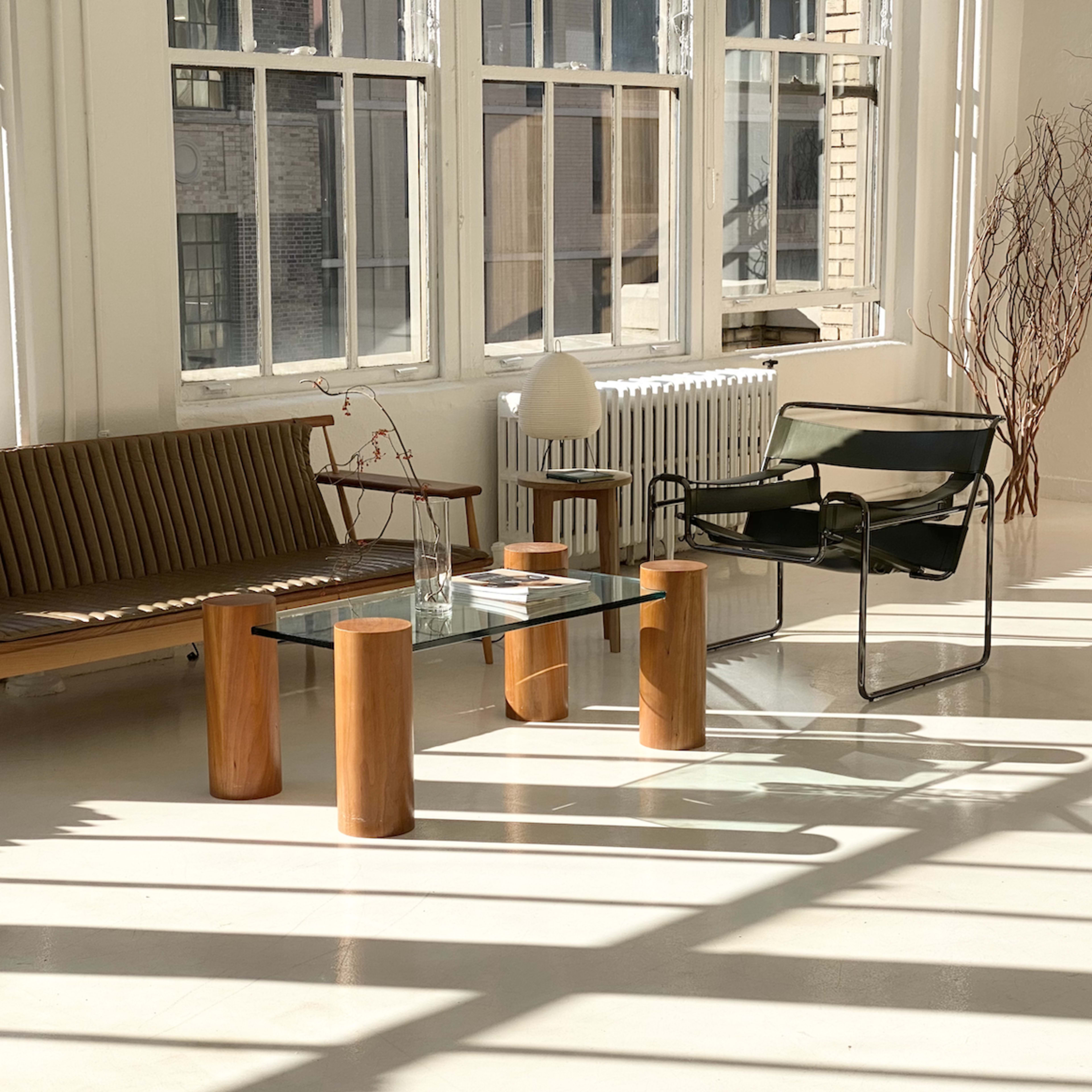 The scene features a bright, minimalist room with a wooden sofa, a glass coffee table supported by cylindrical wooden legs, and a green chair beside a tall wooden structure.
