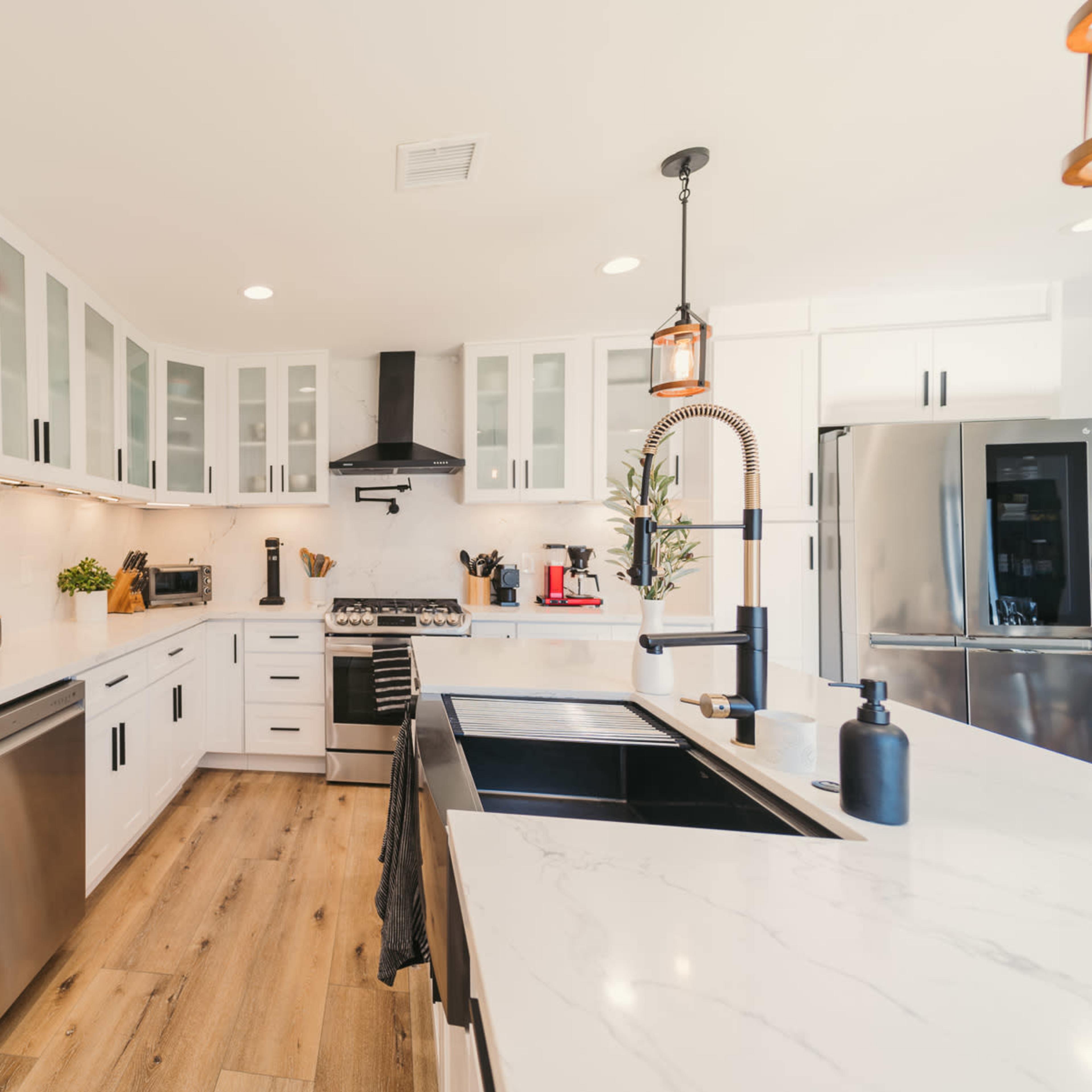 A modern kitchen with white cabinetry, marble countertops, and stainless steel appliances, featuring a large black sink and wooden flooring.
