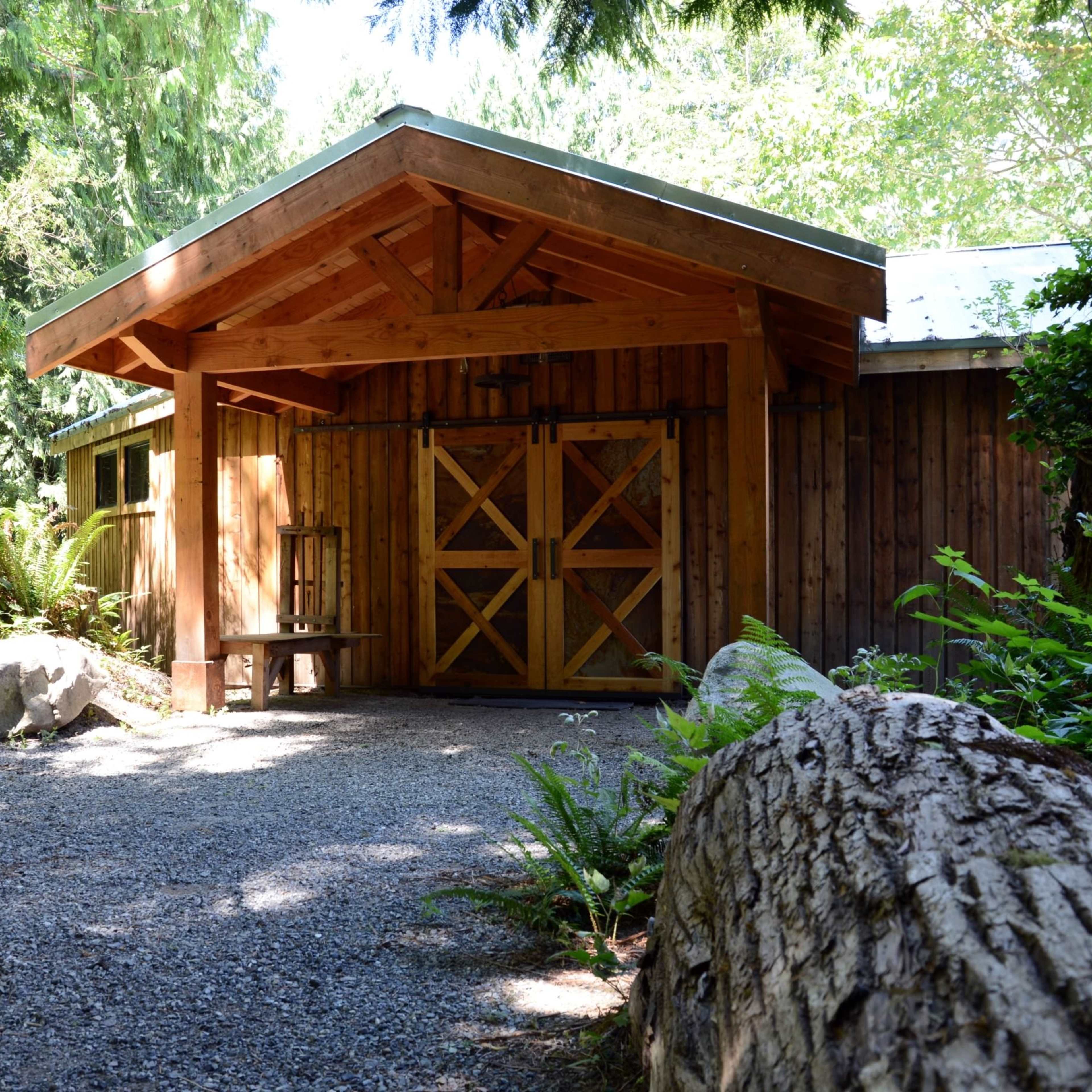 A wooden structure with a peaked roof and double doors is surrounded by trees and ferns along a gravel path.
