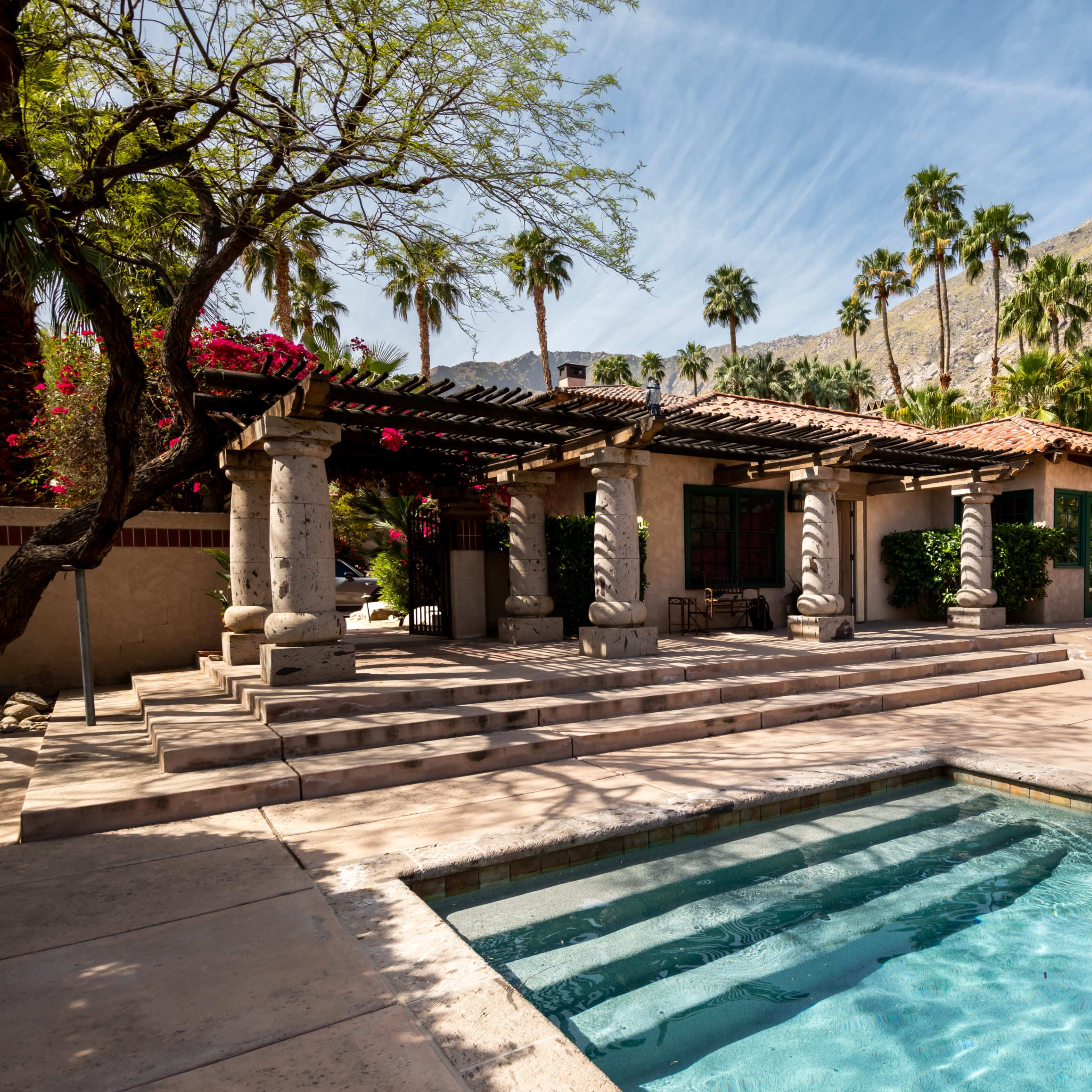 A backyard area featuring a swimming pool, a shaded patio with columns, and surrounding tropical plants and trees against a mountainous backdrop.