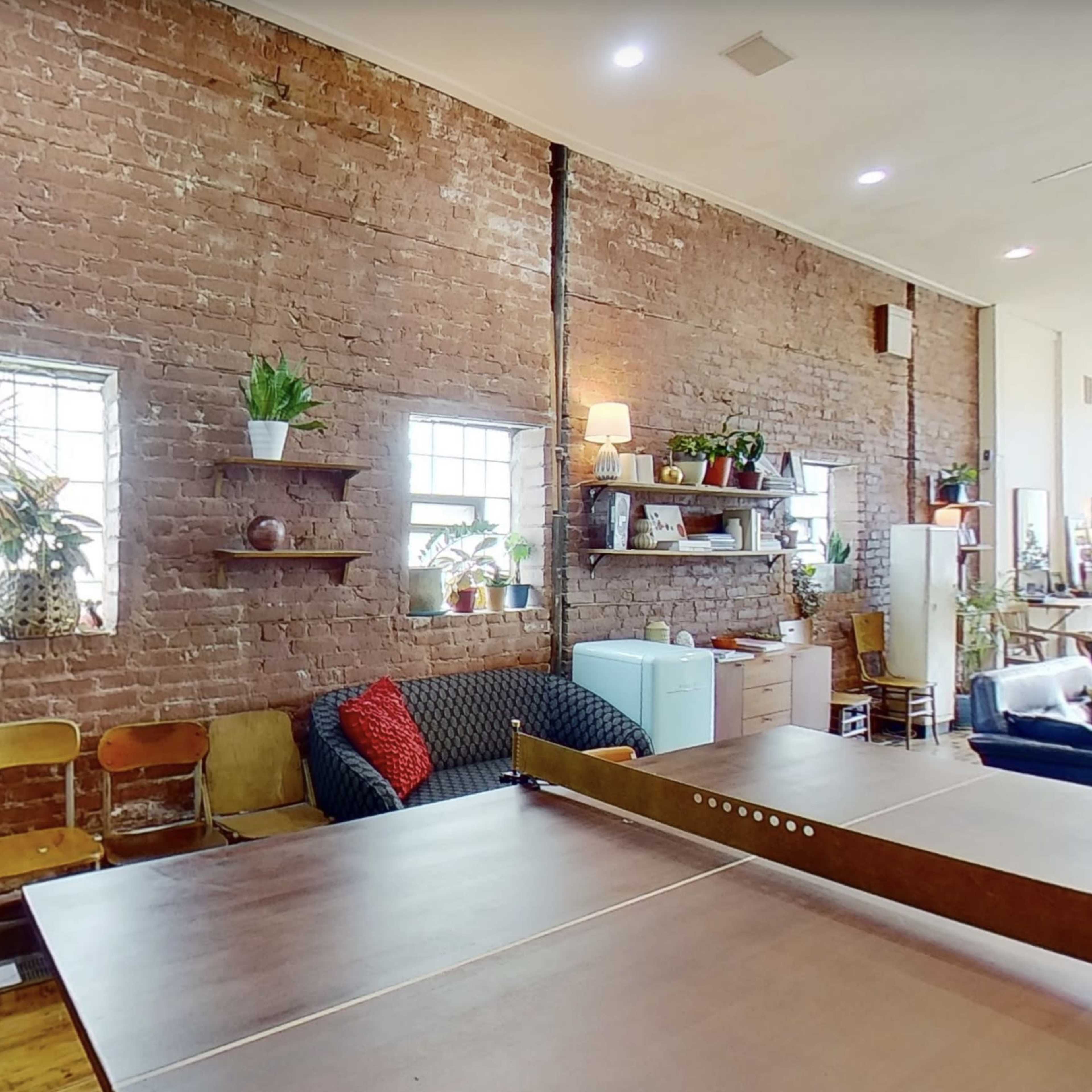 An industrial-style room featuring a brick wall, two windows, shelves with plants, and a ping pong table in the foreground.